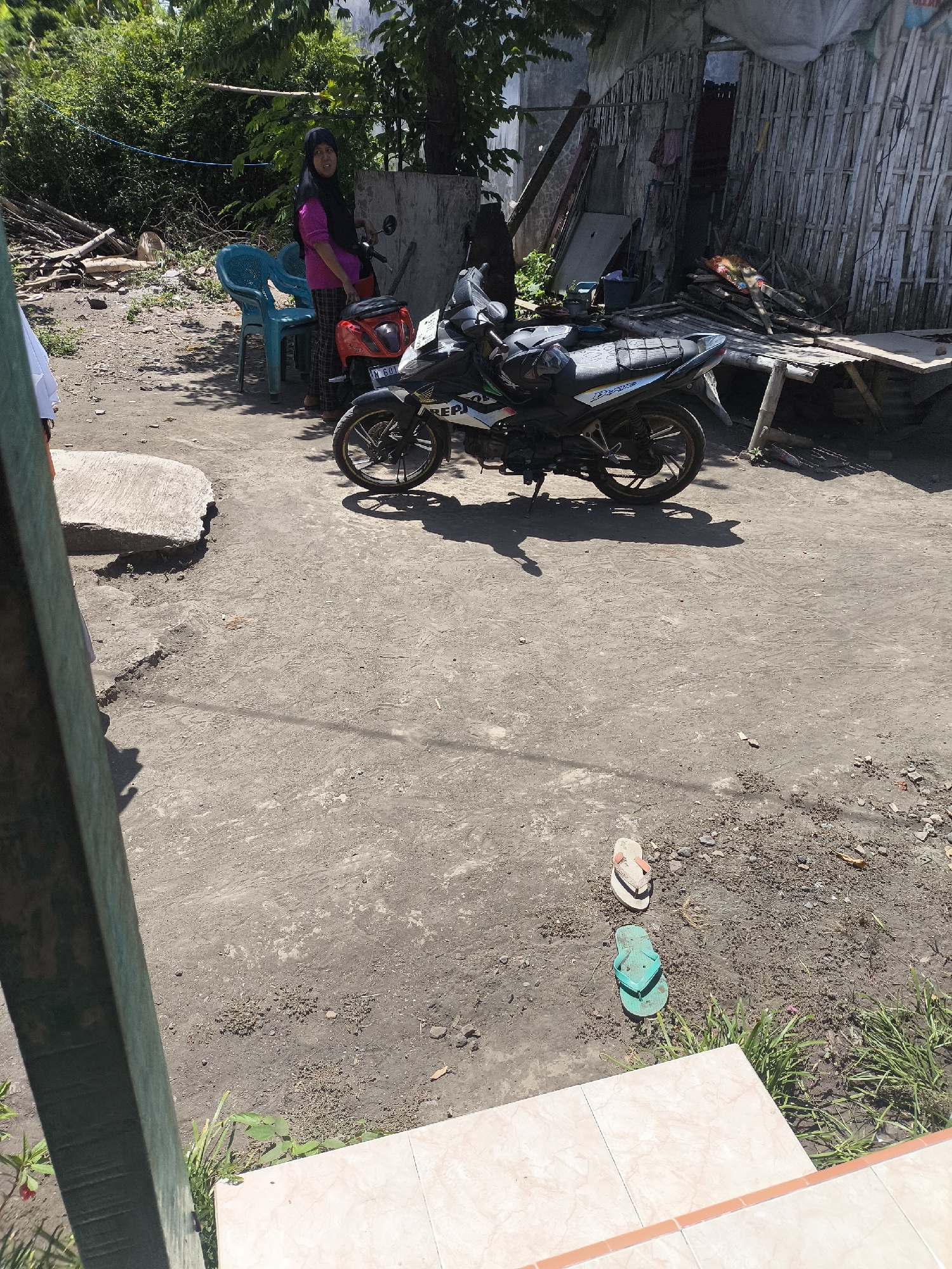 A woman wearing a pink shirt and black hijab stands next to a black and white motorcycle in what appears to be a rural or semi-rural area in Grati, Indonesia. The ground is dirt and there is a cracked concrete slab to the left. In the background, there is a structure made of bamboo and corrugated metal, and a pile of wood. Two blue plastic chairs are to the left of the woman. Further in the foreground, a pair of green flip-flops and a pair of white flip-flops are lying on the ground. The scene is lit by bright sunlight, casting shadows.