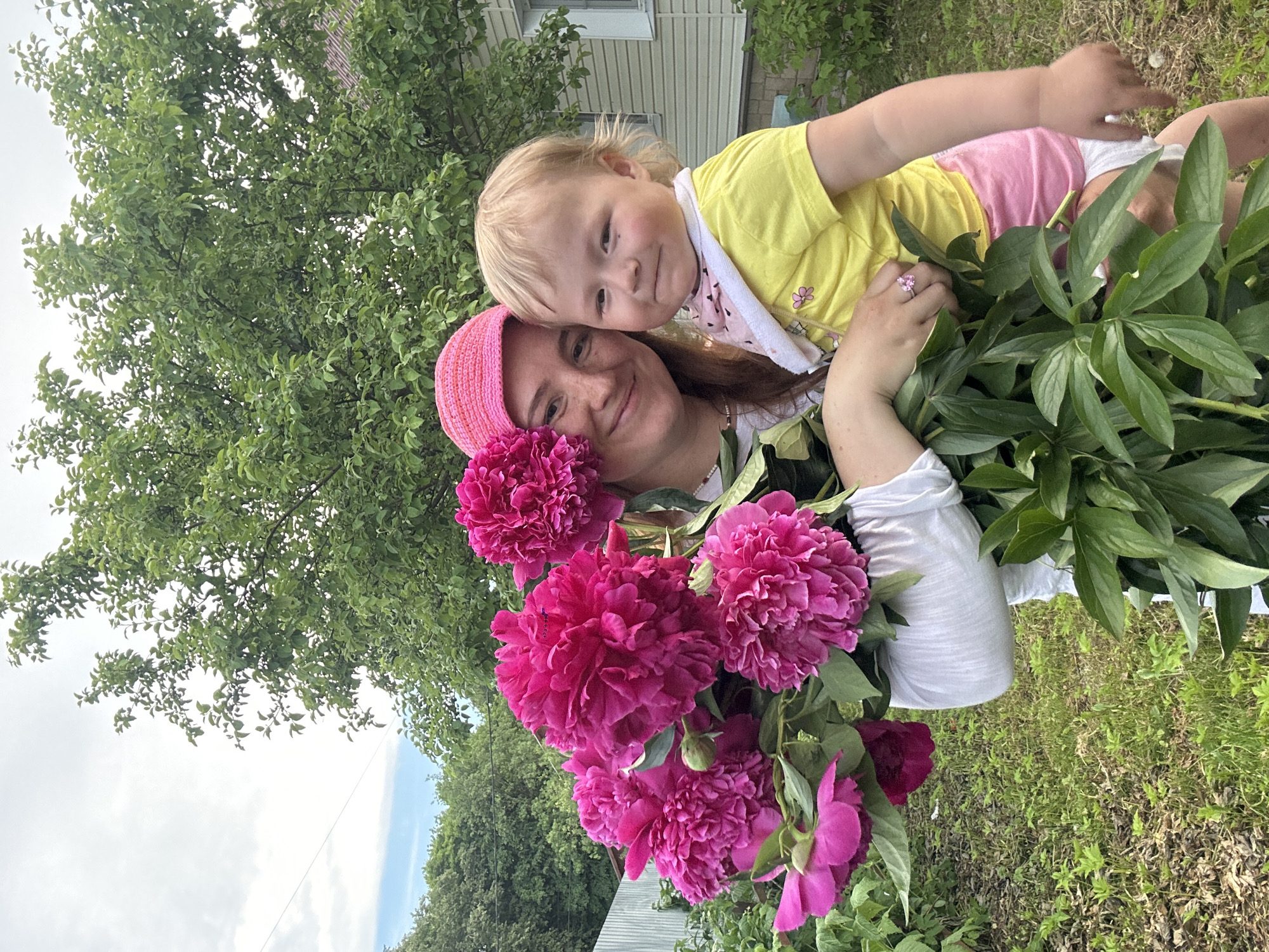 A woman wearing a pink knitted hat holds a toddler while a large bouquet of pink peonies is positioned in front of them. The woman has long brown hair and is smiling at the camera. The toddler, with blonde hair and wearing a yellow t-shirt and pink shorts, looks towards the camera with a slight smile. The background consists of greenery and a light-colored building, suggesting an outdoor setting, possibly a backyard. The image is taken in natural lighting, with a slightly overcast sky. The woman is wearing a white shirt and a necklace. The toddler has a bib around their neck. The peonies are vibrant pink with many petals. The image appears to be taken in Nizhny Novgorod, Russia.