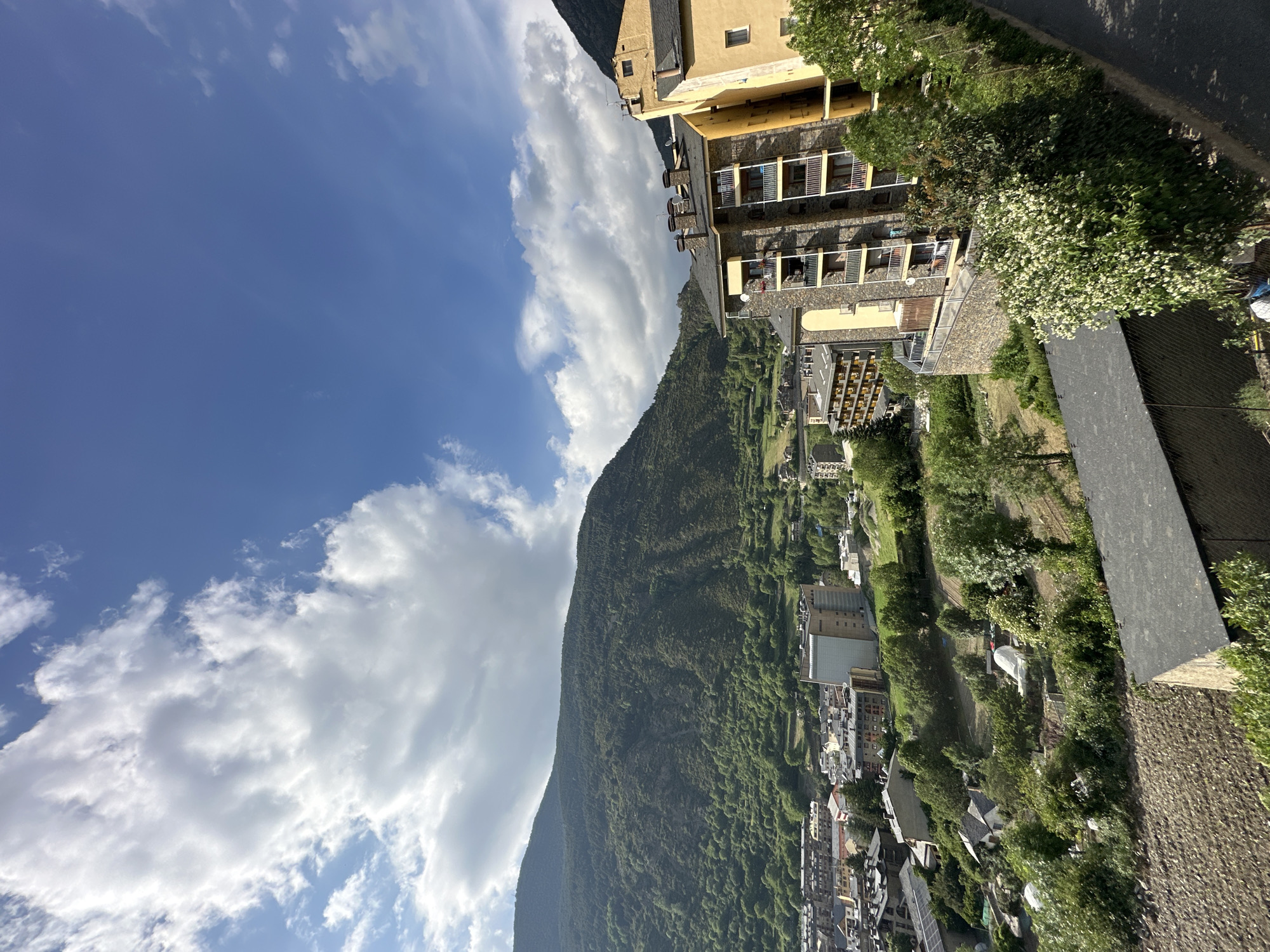 The image presents a panoramic view of a mountainous valley town, identified as Encamp, Andorra, bathed in bright daylight. The scene is oriented to show a clear blue sky, partially adorned with scattered, fluffy white clouds, indicating pleasant weather.
A large, steep mountain dominates the left side of the frame, densely covered with vibrant green trees from base to summit. Its ridgeline forms a prominent diagonal across the image. On the opposing side of the valley, and extending into the foreground, lies the town itself.
In the mid-ground and foreground on the right, numerous buildings are visible, characterized by their European architecture featuring light-colored facades, stone detailing, and dark roofs. A multi-story building with a yellowish exterior and several balconies, complete with chimneys, is prominent in the immediate right foreground. Below and to its left, the town extends with a mix of structures nestled into the valley floor and climbing the lower mountain slopes. Lush green vegetation, including trees and flowering white bushes, is interspersed throughout the town and along the valley. A dark paved road or pathway is visible in the upper right portion of the image.
The strong sunlight suggests it is midday or early afternoon. No human activity or visible text is discernible in the tranquil mountain landscape.