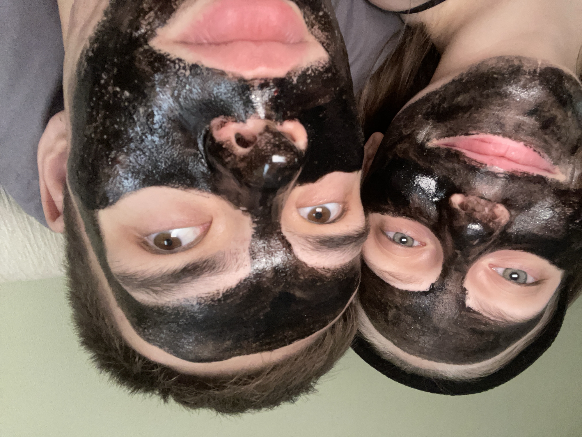 In the image, two individuals are positioned closely together, both wearing black charcoal face masks that obscure much of their facial features. The person on the left appears to have light-colored eyes and is wearing a black headband. The person on the right has brown eyes and dark hair. The lighting suggests an indoor setting, possibly a home, with a plain background. There is no identifiable information within the image to suggest a specific location in Ufa, Russia, or any other city. The image focuses on the faces of the individuals and their masks, with minimal environmental context.