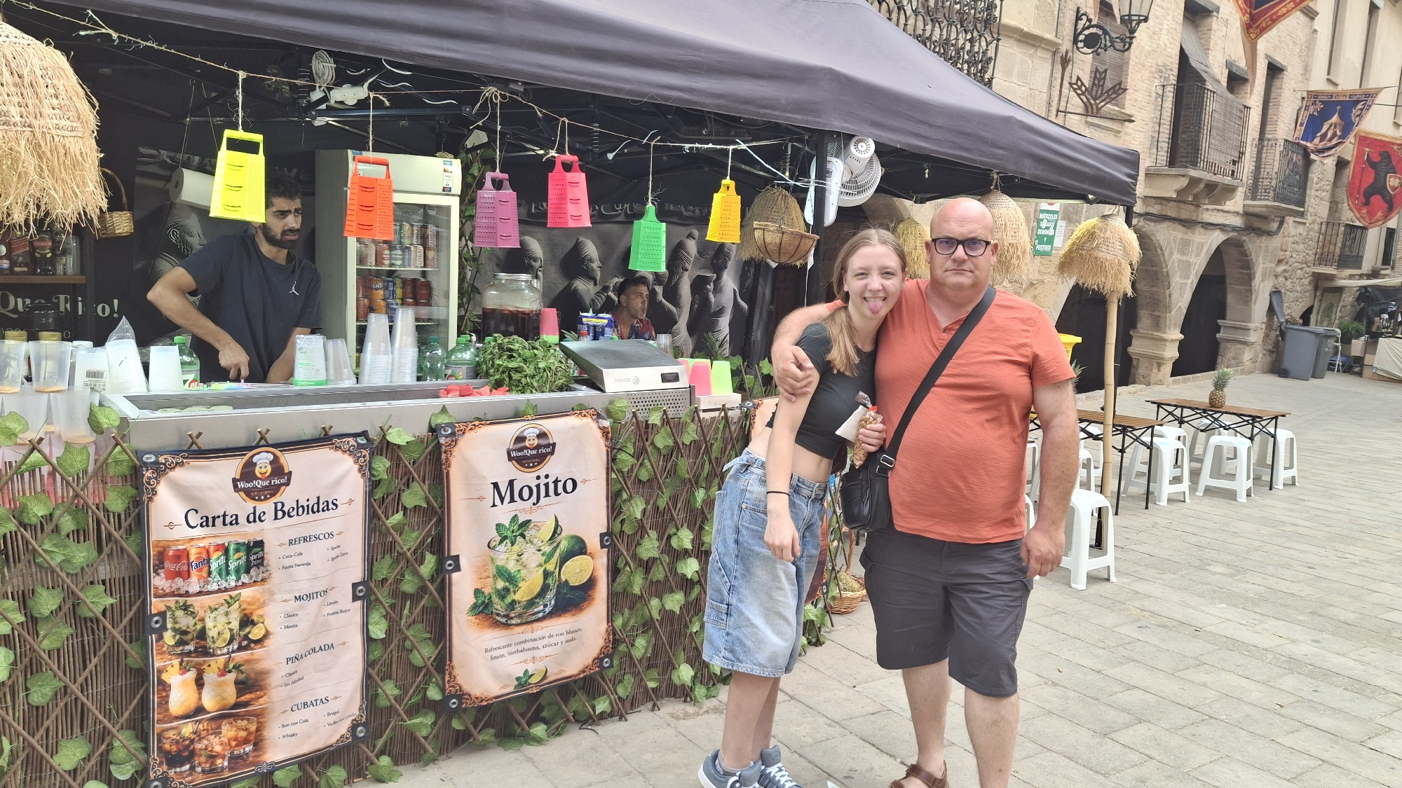 In La Fresneda, Spain, an outdoor beverage stand is depicted. A man with a beard, wearing a dark t-shirt, is visible behind the counter, preparing drinks. Hanging from the awning above are several brightly colored grater-shaped decorations in yellow, orange, pink, and green. A young woman, wearing a black crop top and denim shorts, stands to the left of a man in an orange t-shirt and dark shorts. They are posing for a photograph in front of the stand. The stand displays a menu board advertising drinks like "Carta de Bebidas," "Refrescos," "Mojitos," "Piña Colada," and "Cubatas." In the background, there are outdoor seating areas with tables and stools, as well as a stone building with arches. The ground is paved with stone tiles.