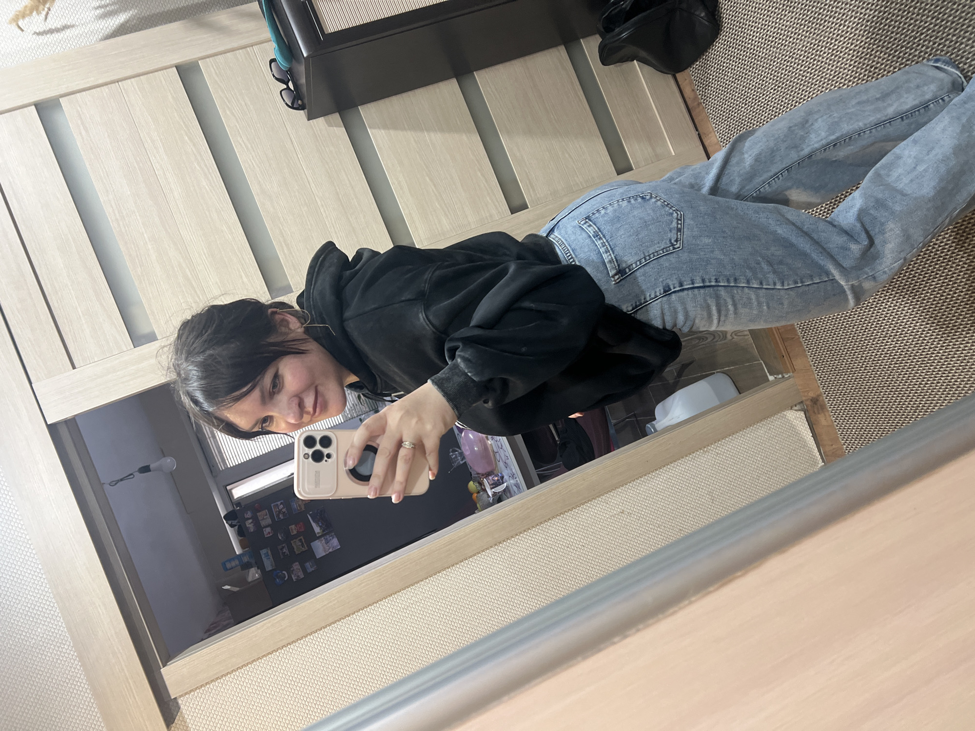 A person is taking a selfie in a mirror. The person is wearing a black hoodie and light blue jeans. A ring is visible on their left ring finger. The reflection in the mirror reveals a kitchen with a refrigerator covered in magnets and notes. A lightbulb hangs from the ceiling. The surrounding environment includes a wardrobe with a dark-colored cabinet on top, a brown woven rug, and what appears to be an interior door. The location is implied to be in Noviy, Russia.