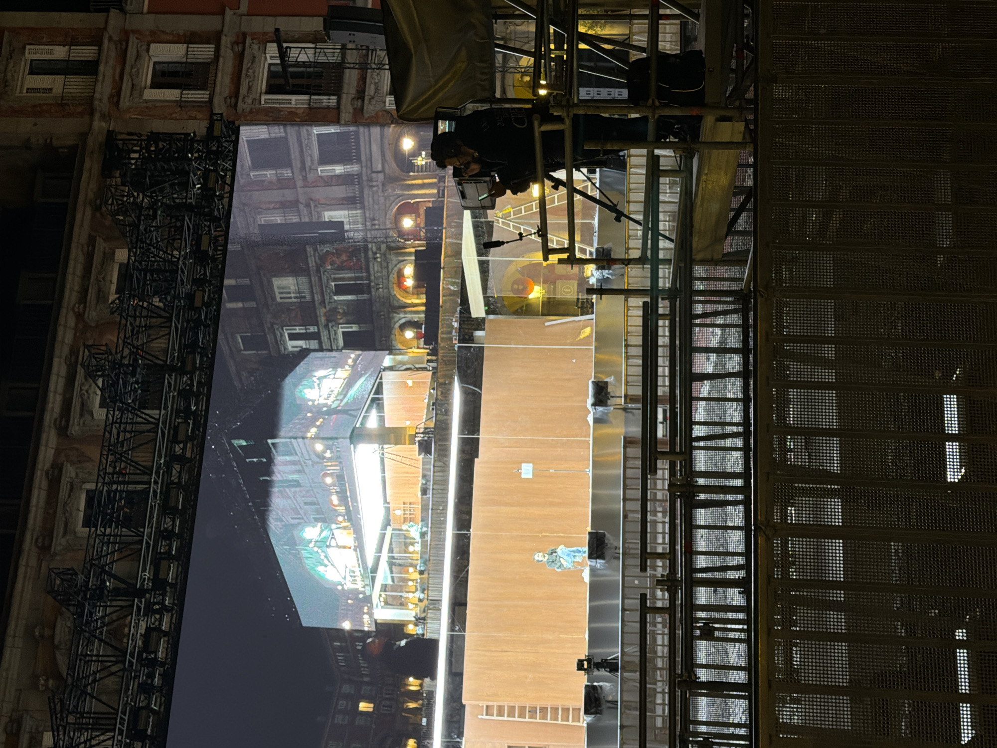 The image shows a scene at night in Madrid, Spain, with a large screen displaying a building facade reflecting the actual building on the left. The screen is part of a stage setup, with scaffolding visible on the right and supporting a person operating equipment at the top. The ground in front of the screen appears to be a performance space with a ladder, and the setting seems to be a building exterior in the heart of Madrid's historic district.