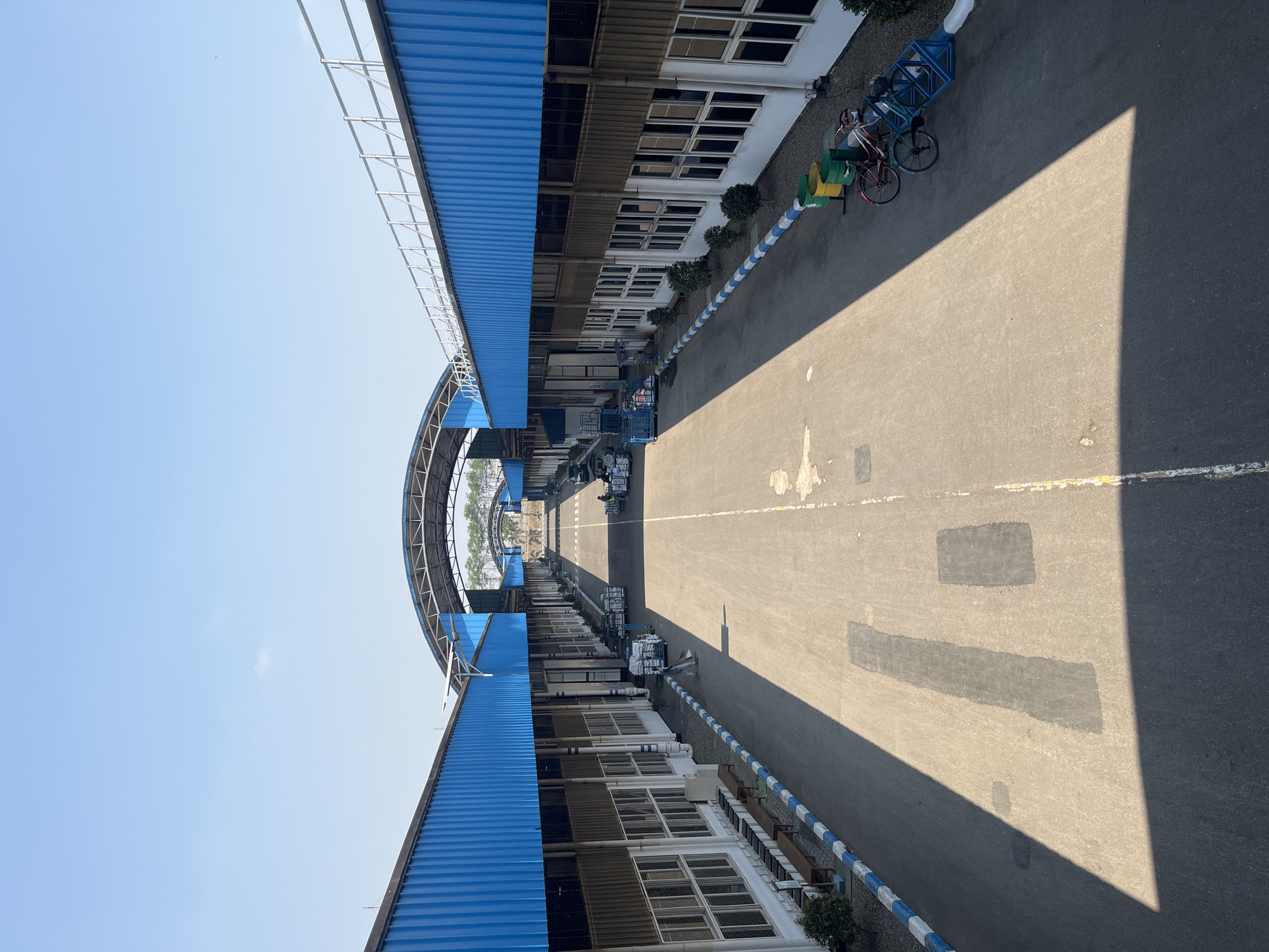 This is a daylight, eye-level, long-shot, wide-angle photograph of an industrial complex with an arched walkway in Pelang, Indonesia. The scene is dominated by long, single-story buildings with bright blue corrugated metal roofs. The buildings have a uniform facade with rows of windows. The ground is paved asphalt, with a clear demarcation line down the center of the walkway. The walkway is lined with blue and white striped bollards.
In the foreground, the walkway is mostly empty, with a long shadow cast from an unseen object to the left. There are some patches of darker asphalt on the ground, possibly indicating repairs.
Further down the walkway, there are several objects and a few people. On the left side, there are benches placed outside the building. On the right side, there are two bicycles parked next to some green and yellow cylindrical bins. Further along, there are several industrial carts filled with white sacks, and a few individuals are seen pushing or attending to them.
The arched walkway extends into the distance, where it connects to similar structures, creating a repeating architectural pattern. Trees are visible in the background beyond the complex. The sky is clear and blue, indicating a sunny day.