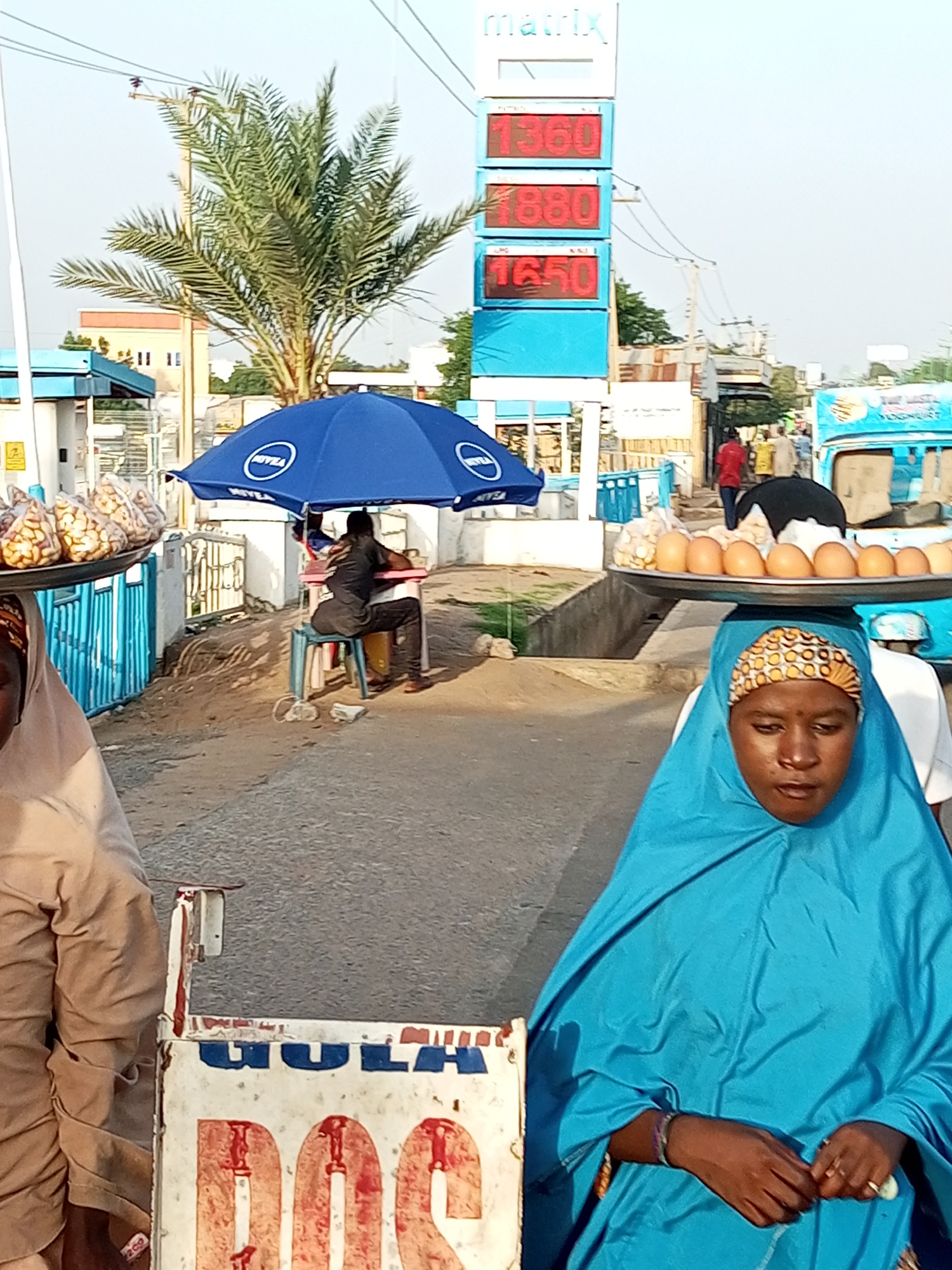 In Garko, Nigeria, a street scene unfolds with a focus on a price display board for a fuel station. The board shows prices of 1360, 1880, and 1650 in red digital numbers against a blue background, with the "Matrix" logo at the top. In the foreground, two women are depicted. The woman on the right wears a bright blue hijab and carries a tray of eggs on her head, while the woman on the left, partially visible, wears a beige head covering and also carries a tray of food items. Behind them, a person sits under a blue umbrella advertising "Nivea" brand, appearing to be a vendor or attendant. Several people, including some in red and beige clothing, are visible walking in the background towards a blue and white vehicle. A palm tree stands to the left of the fuel price board, and the ground is a mix of asphalt and dirt. A white cart with red and blue markings, displaying "GULA DOS," is positioned in the lower left corner. The environment suggests a bustling outdoor marketplace or roadside area.
