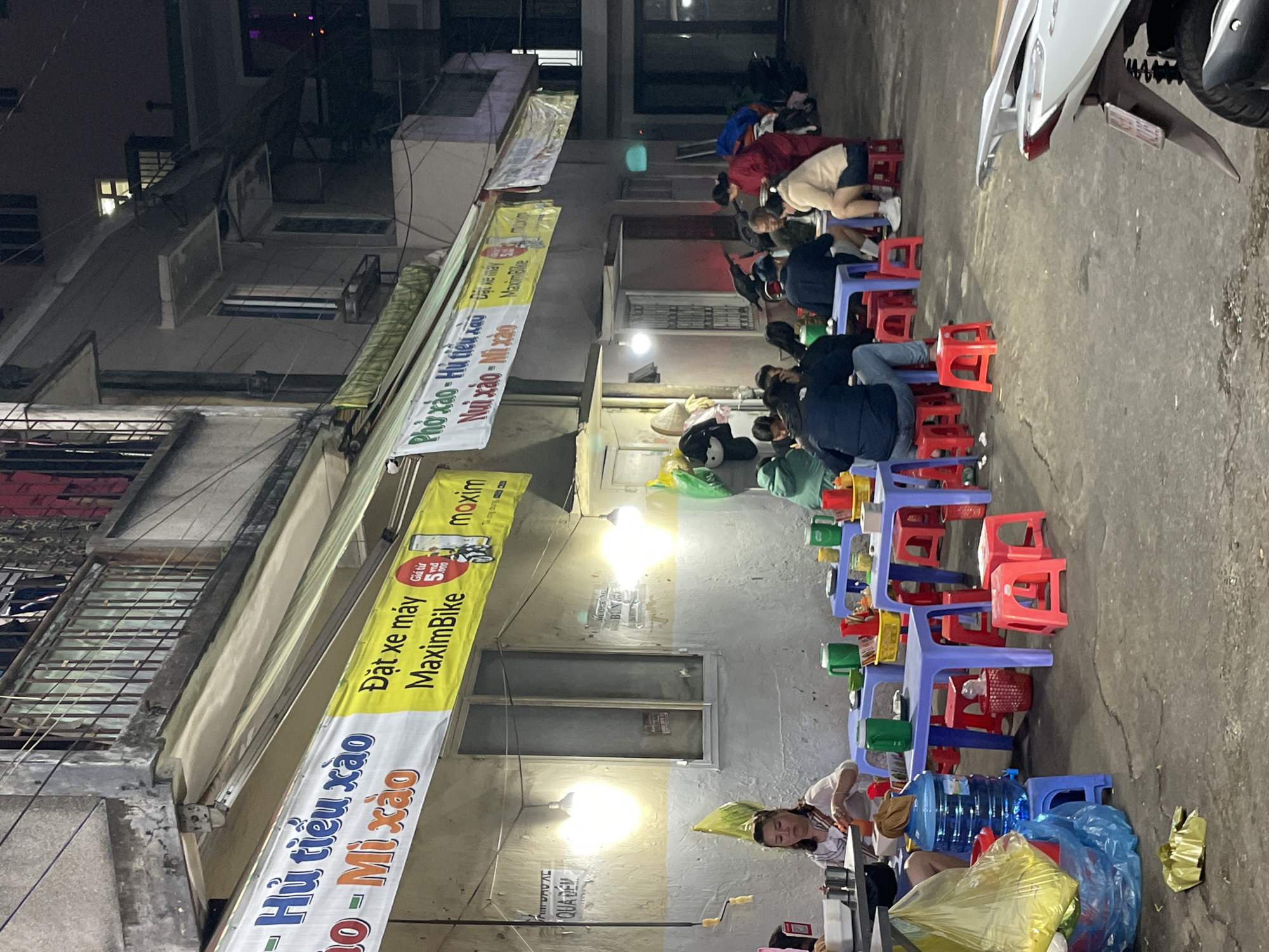 The image depicts an outdoor dining area at night in Da Lat, Vietnam, likely a street food establishment. Several small, brightly colored plastic tables and stools are set up on a concrete ground. People, dressed in casual attire, are seated at the tables, some eating and others conversing. The scene is illuminated by artificial lights, including overhead bulbs and lights from within the buildings. Banners advertising food items like "Phở xào - Hủ tiếu xào" (stir-fried noodles - stir-fried rice noodle soup) and "Nui xào - Mì xào" (stir-fried macaroni - stir-fried noodles) are visible, along with a banner for "MaximBike" which appears to be a delivery service. There are also large water cooler jugs and various food containers and utensils on the tables. In the background, a multi-story building with balconies and laundry hanging out can be seen. A white scooter is parked in the foreground on the right. The overall atmosphere suggests a bustling, informal eatery.