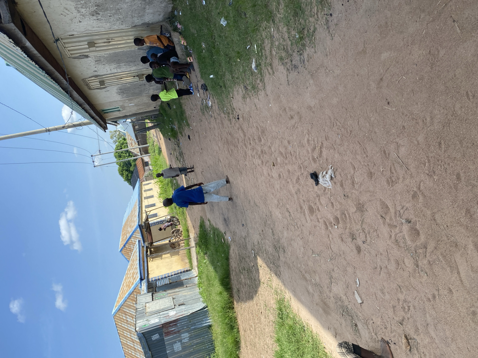 A group of young boys are gathered outside a building in Jauro Yinu, Nigeria. The scene appears to be a dusty, sandy area with patches of grass and a few buildings with corrugated metal roofs. The boys are wearing casual clothing, with some in t-shirts and shorts. One boy in a bright green shirt stands out from the group. In the background, a few more individuals are visible, including a man in a blue shirt standing near a cart. The sky is clear with some white clouds, suggesting it is daytime. The overall impression is of a typical day in a rural or semi-urban setting in Nigeria.