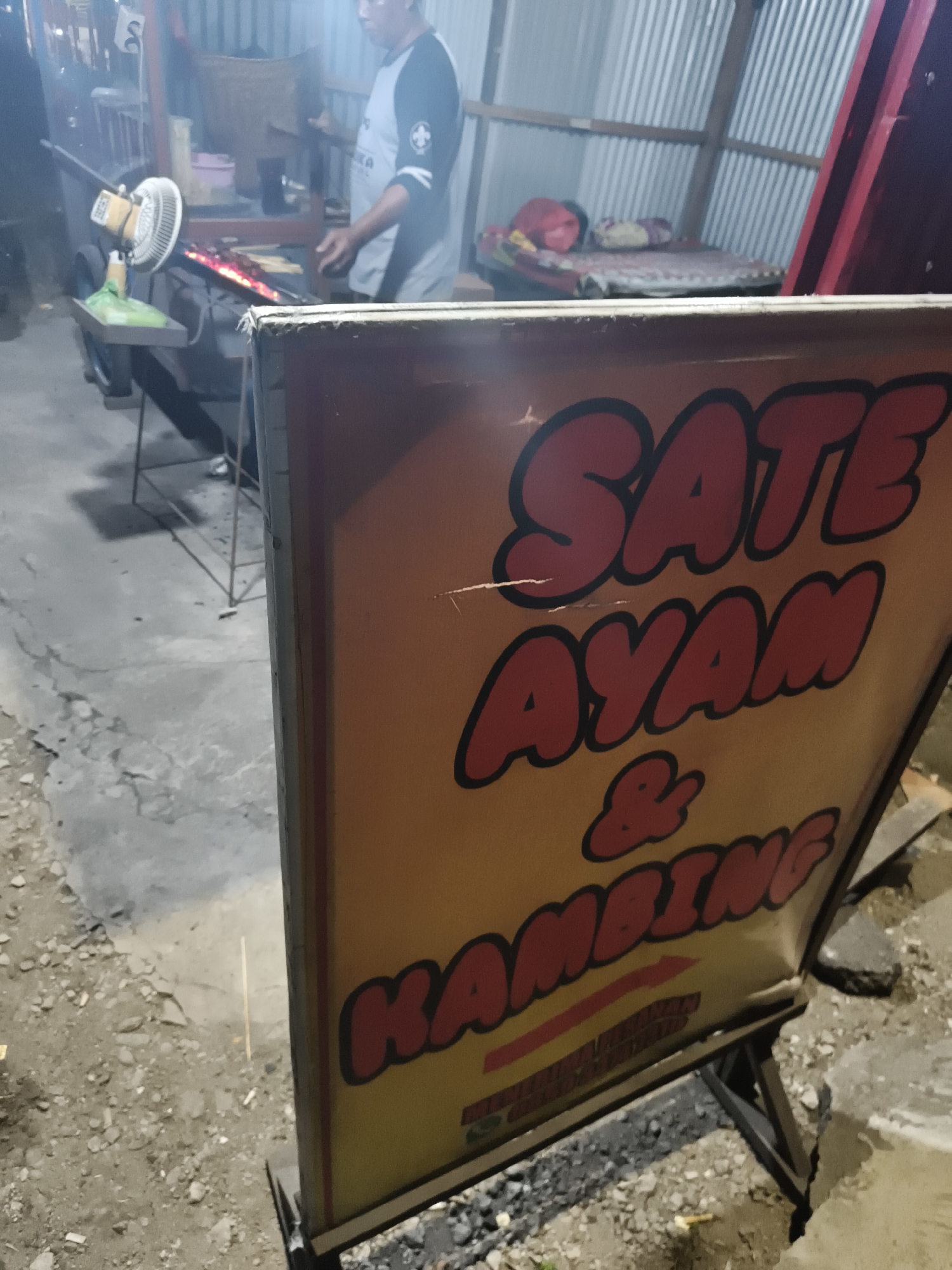 In the city of Sidorejo, Indonesia, at night, a food stall sign is prominently displayed in the foreground. The sign has a bright yellow background with large, red, bubble-style lettering outlined in black. The text reads "SATE AYAM & KAMBING," indicating that the stall sells chicken and goat satay. Below this, there appears to be additional text or a logo, but it is too blurry to decipher. The sign is tilted and supported by a dark metal stand. In the background, out of focus, a person wearing a gray and black t-shirt is tending to a grill where satay skewers are being cooked over an open flame. To the left of the grill, a yellow fan is positioned on a stand. Further in the background, a simple sleeping area with a mattress and pillows is visible under a corrugated metal roof. The ground is a mix of dirt and gravel. The overall scene suggests a casual, open-air food vending operation.
