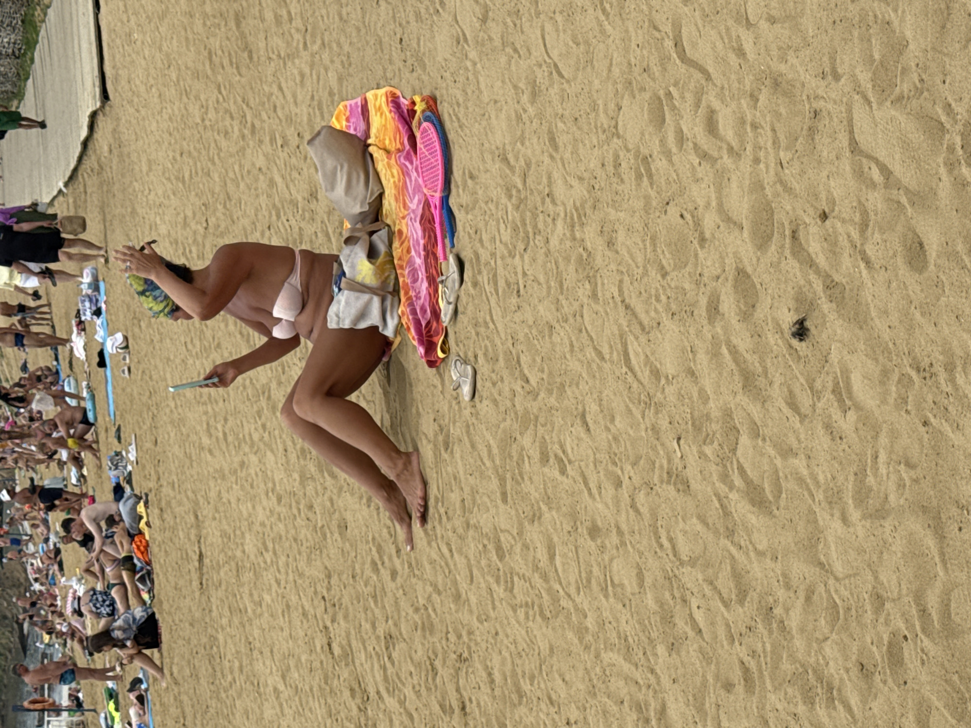 The image depicts a crowded beach scene in Yarovoye, Russia. In the foreground, a woman is sitting on a colorful towel, wearing a light pink bikini top and a headscarf. She is holding a smartphone in her right hand and appears to be adjusting her headscarf with her left hand. Beside her, on the towel, are a pair of flip-flops, a tan tote bag, and a pink and blue beach racket. In the background, numerous people are spread across the sandy beach, some sunbathing, others walking. The beach appears to be a popular recreational area, with a wooden walkway visible in the upper right corner. The sand is light brown and textured with footprints.