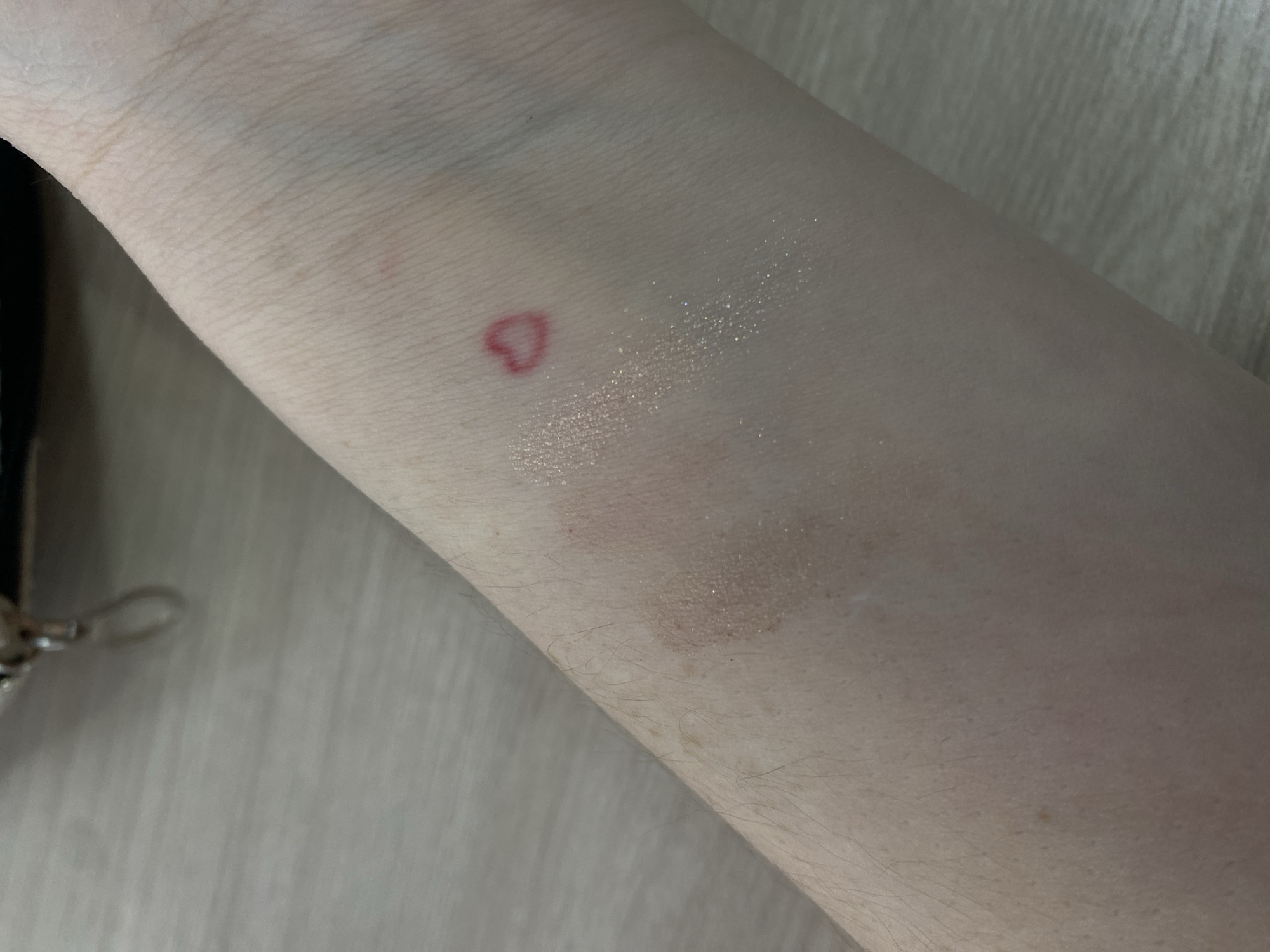 The image shows a forearm, likely of a person, resting on a light-colored wooden surface. There is a red heart shape drawn or marked on the skin. There are also areas of what appear to be shimmery, light-colored cosmetics swatched near the heart. A small part of what looks like a black bag with a silver clasp is visible on the left side of the frame. There is no indication of location. The city of Chaikovsky, Russia is not visible in this image.