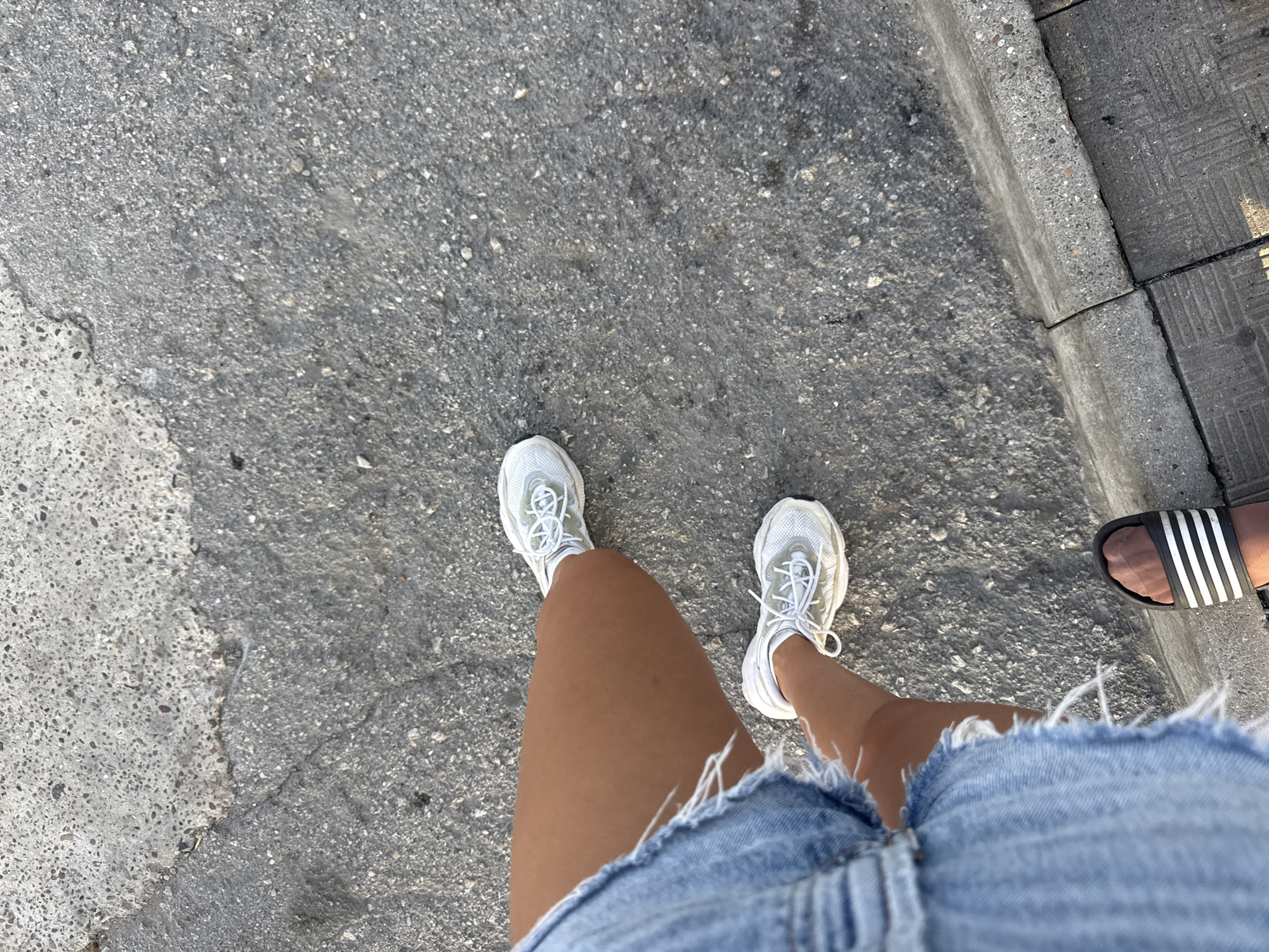 The image shows a close-up, high-angle view of a person's legs and feet walking on a grey asphalt surface. The person is wearing a distressed denim skirt and white sneakers. One foot is also visible wearing a black and white striped slide sandal. The asphalt appears worn and cracked in places. The scene suggests an outdoor urban environment, possibly a sidewalk or street. The specific location in Cimișlia, Moldova, cannot be determined from the visual information present.