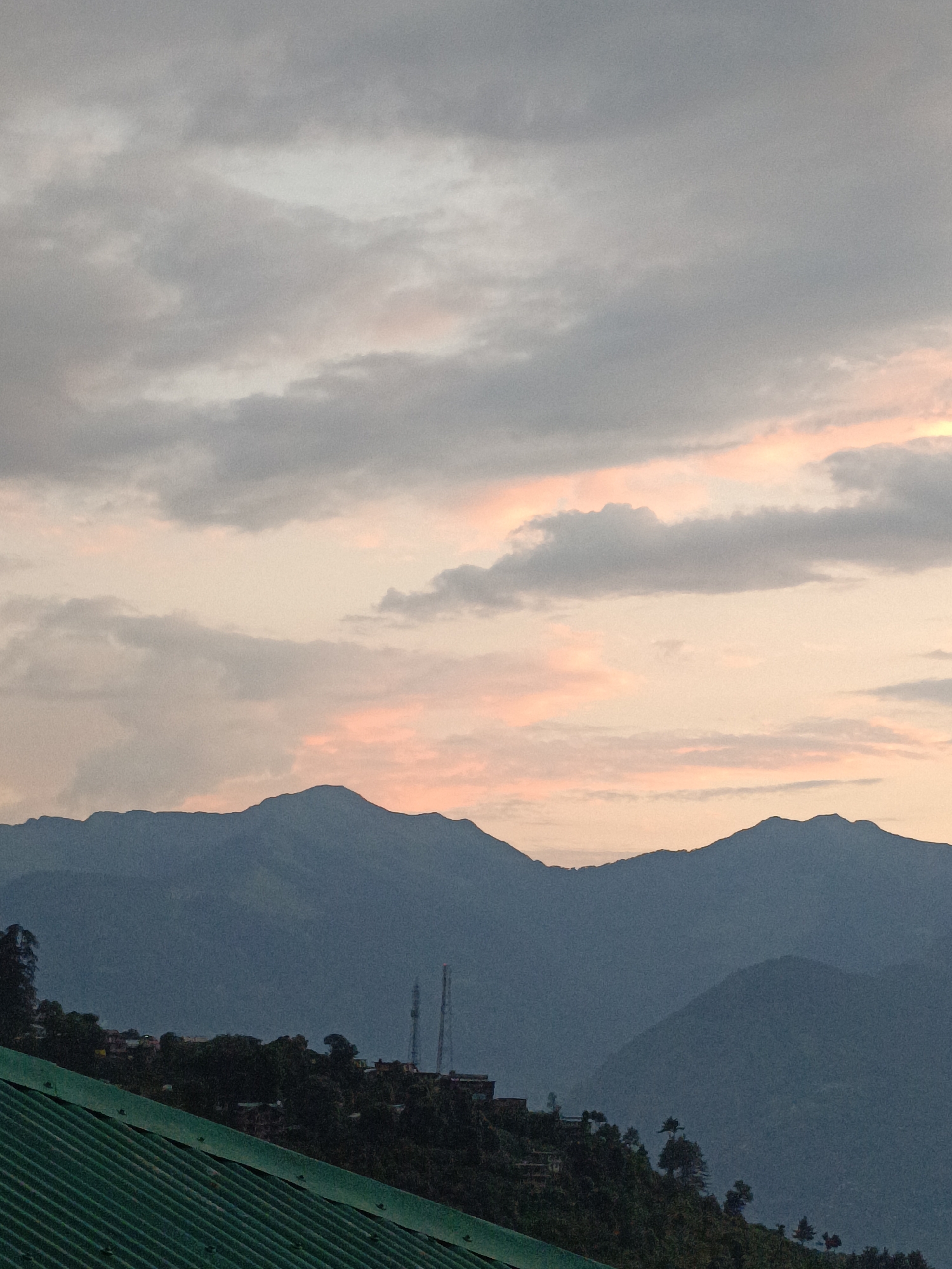 The image depicts a mountainous landscape at dusk, likely in or near Damadi, India, given the context. The foreground is dominated by a portion of a green corrugated metal roof, angled from the bottom left corner upwards and towards the right. Beyond the roof, a densely forested hillside is visible, dotted with scattered buildings, suggesting a settlement. Two tall, slender telecommunication towers rise above the trees in the mid-ground, their metallic structures silhouetted against the sky. The background consists of several layers of majestic, dark blue mountain ranges. The sky above the mountains is a gradient of soft, muted colors, with wispy clouds tinged with pale pink and orange, indicating the setting sun. The overall impression is one of natural beauty and a peaceful, rural setting.