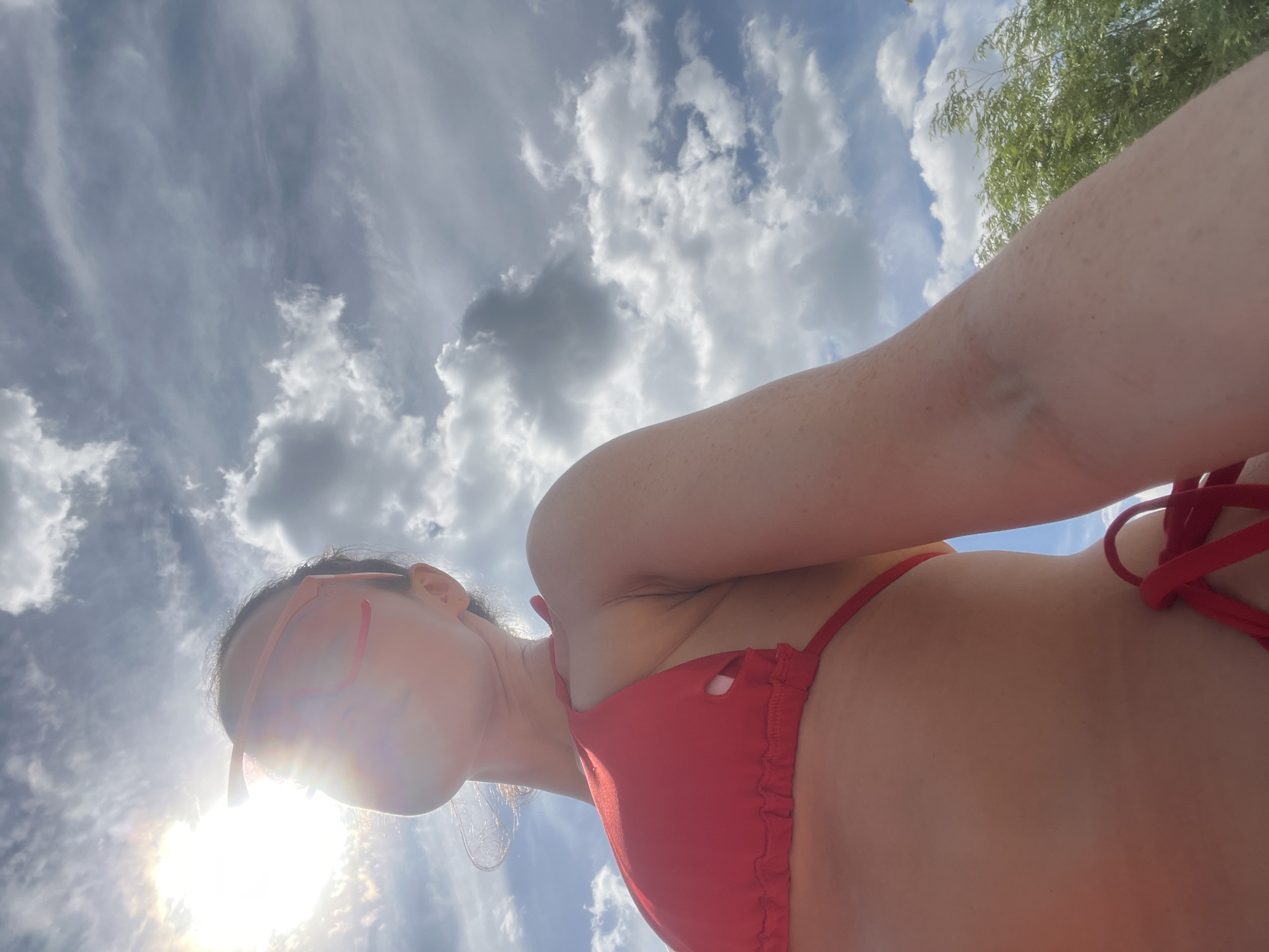 A low-angle, close-up selfie shows a woman wearing a red bikini and orange sunglasses against a sky filled with dramatic clouds. The sun is bright and partially obscured by clouds, creating lens flare. The woman's face is mostly hidden by her sunglasses and the glare. The background consists of a blue sky with white and gray clouds, and a glimpse of green foliage on the right side. The image appears to have been taken outdoors on a sunny day. The location in Kazan, Russia is not discernible from the image.