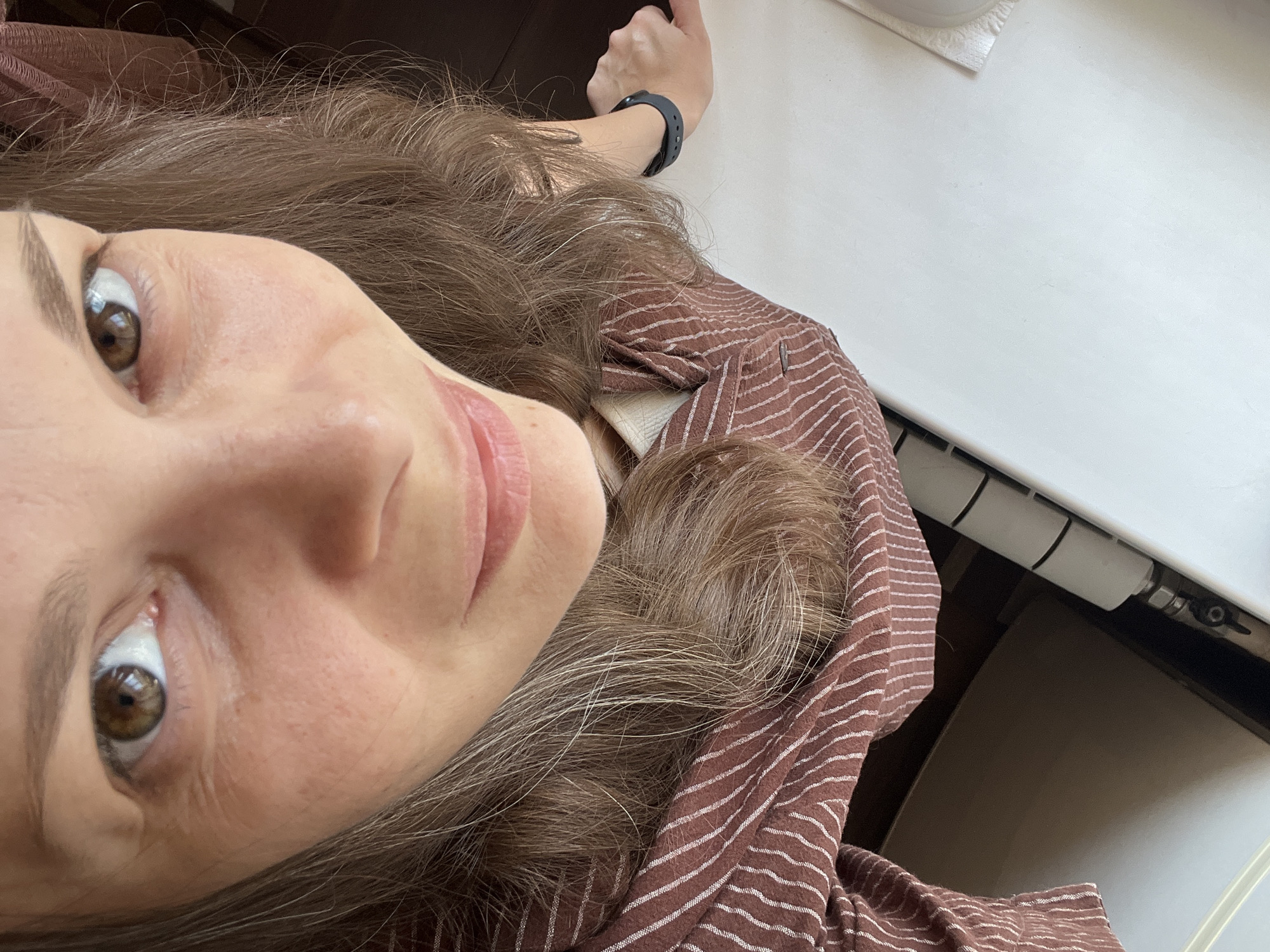 A woman with brown hair and hazel eyes is looking down at the camera. She is wearing a brown and white striped shirt. Her left hand is visible, wearing a black watch. She is sitting at a white table. The background shows a dark wooden surface and a white object with black stripes. The image is taken from a high angle, looking down at the woman. There is no indication of the location in Tolyatti, Russia.