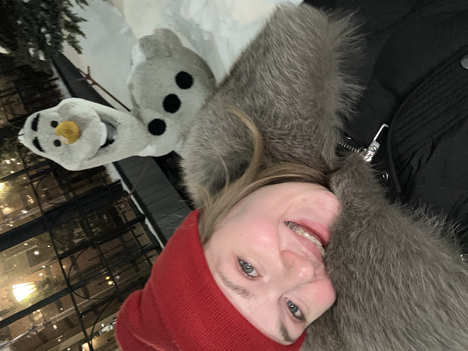 A woman in a red beanie and a fur-trimmed coat smiles at the camera. She has braces. Next to her, a plush toy of Olaf from Frozen, with a carrot nose, is followed by a gray bunny with black eyes. In the background, there's a glimpse of snow-covered ground and greenery. Additionally, some lights from buildings can be seen.