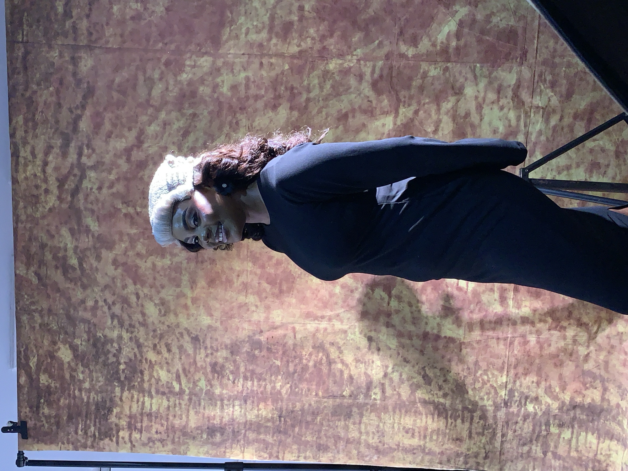 A full-body, side-profile shot shows a Black woman with dark, curly hair and fair skin, wearing a black, long-sleeved dress and a white wig. She is standing in front of a textured, brown and yellow backdrop, possibly a painted canvas, that appears to be set up in a studio or event space. The woman is smiling and looking to her left. A portion of a dark metal stand is visible on the right side of the frame, suggesting the backdrop is suspended or supported. The image is taken indoors, with artificial lighting that casts shadows on the backdrop. The location is not specified in the image or provided context.