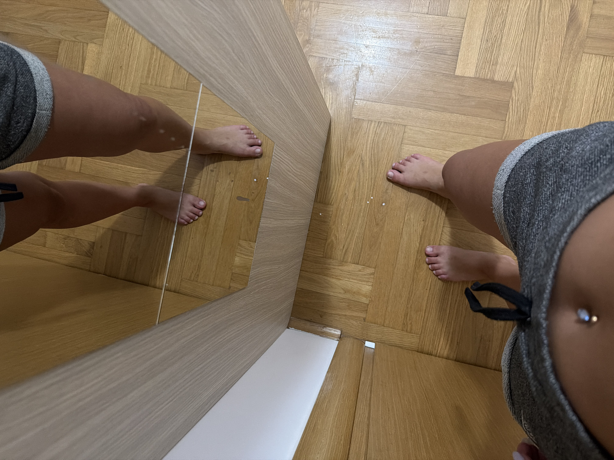 The image captures a low-angle, full-body shot of a person standing barefoot on a wooden parquet floor in what appears to be an indoor residential setting. The person is wearing grey shorts with a drawstring and a visible navel piercing. The individual's legs and feet are prominent in the foreground and reflected in a mirror that occupies the upper half of the image. The mirror reveals the person's thighs and knees from a slightly different perspective, also showing the wooden floor. The environment is sparsely decorated, with a light-colored wall or cabinet visible to the left. The lighting is moderate, creating some subtle shadows. There is no indication of the city of Budva, Montenegro, or any specific urban elements within the image.