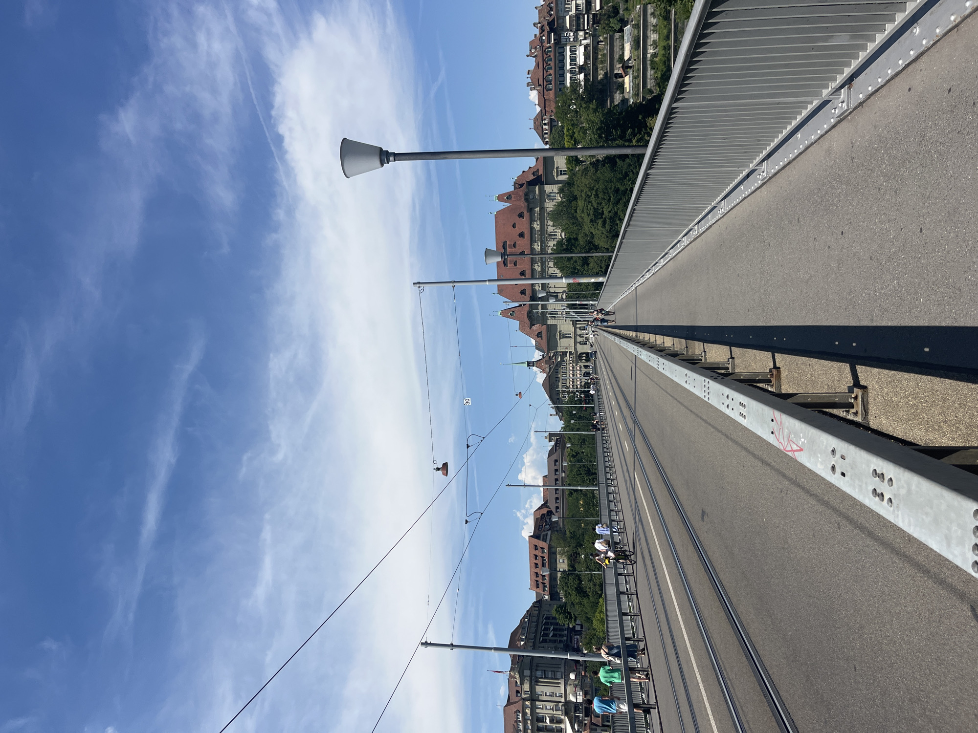 A wide shot captures a perspective from a bridge in Bern, Switzerland, looking forward along its length. The bridge, which appears to be a thoroughfare for vehicles and pedestrians, features tram tracks running down its center. On the left side of the bridge, several individuals are seen walking and cycling. The right side of the bridge is bordered by a metal railing. Beyond the bridge, the cityscape of Bern unfolds with numerous buildings, some of which have red-tiled roofs, and lush green trees. The sky above is a clear blue with scattered white clouds, indicating a pleasant day. Overhead, power lines for the trams are visible, along with lampposts positioned along the bridge.