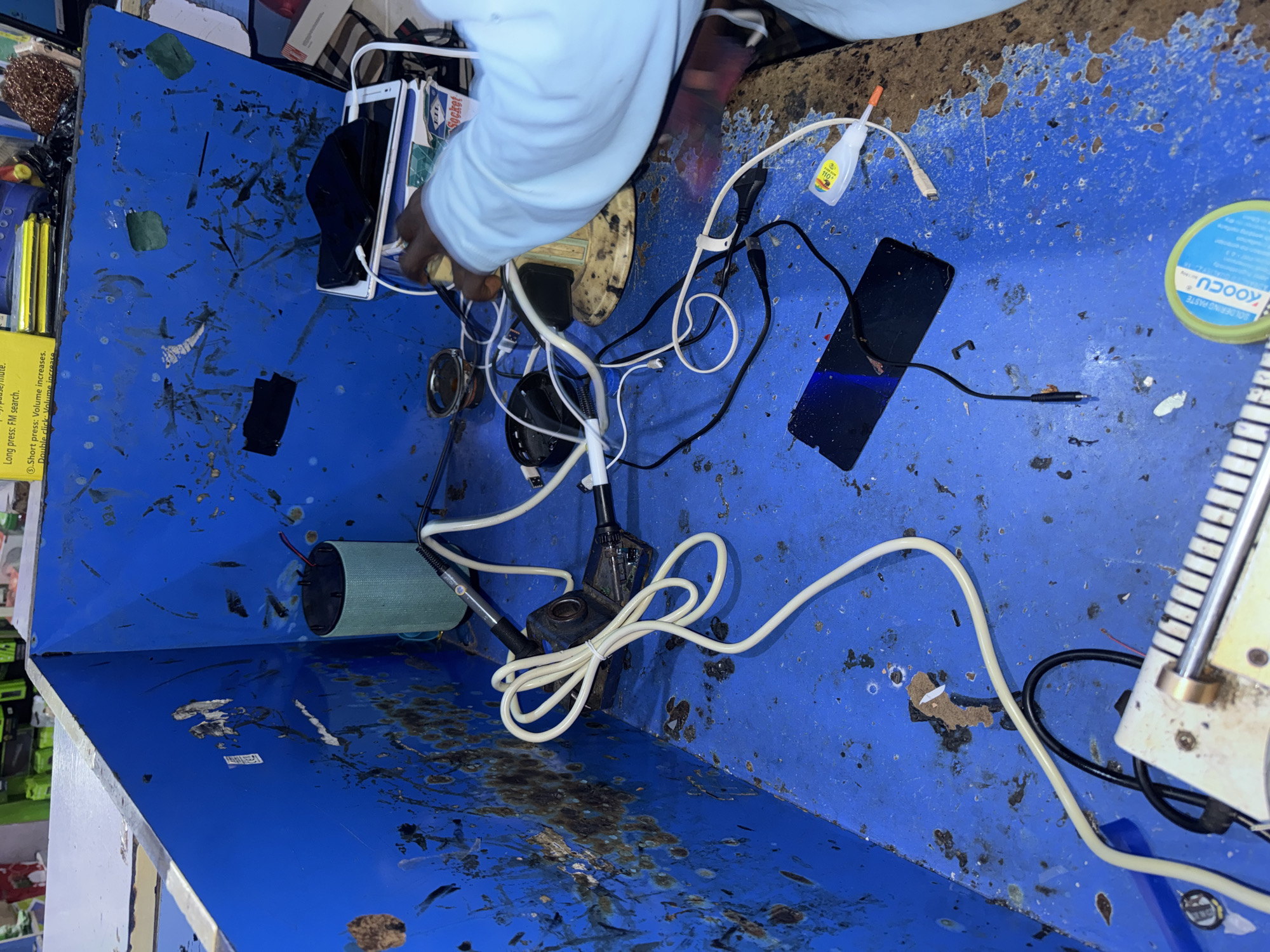 The image depicts a cluttered, blue metal bin filled with various electronic components and wires. A person, wearing a light blue long-sleeved shirt, is partially visible, holding a white and black smartphone and appearing to be engaged in some sort of repair or tinkering.
Among the objects are several cables, including a white coiled cable, black USB cables, and a power cord. A smartphone lies face down on the blue surface, with a black charging cable plugged into it. A small bottle of what appears to be glue is also present, along with a round container labeled "KOOCU SOLDERING PASTE." A cylindrical object, possibly a speaker or a component for a device, is positioned on the left side of the bin. The bin itself is the setting, appearing to be a workspace or storage area for discarded electronics. The blue surface of the bin is marked with rust and grime, suggesting it might be outdoors or in a workshop environment.
The visible text includes instructions for a device: "Long press: FM search.", ".Short press: Volume increases.", and "Double click: Volume increase". The brand name "KOOCU" and "Socket" are also legible.
Given the location context of Jalingo, Nigeria, this scene likely represents a repairman or electronics enthusiast working with salvaged or broken devices. The overall impression is one of a hands-on activity, possibly involving DIY electronics repair or recycling. The time of day and weather are not discernible from the image.