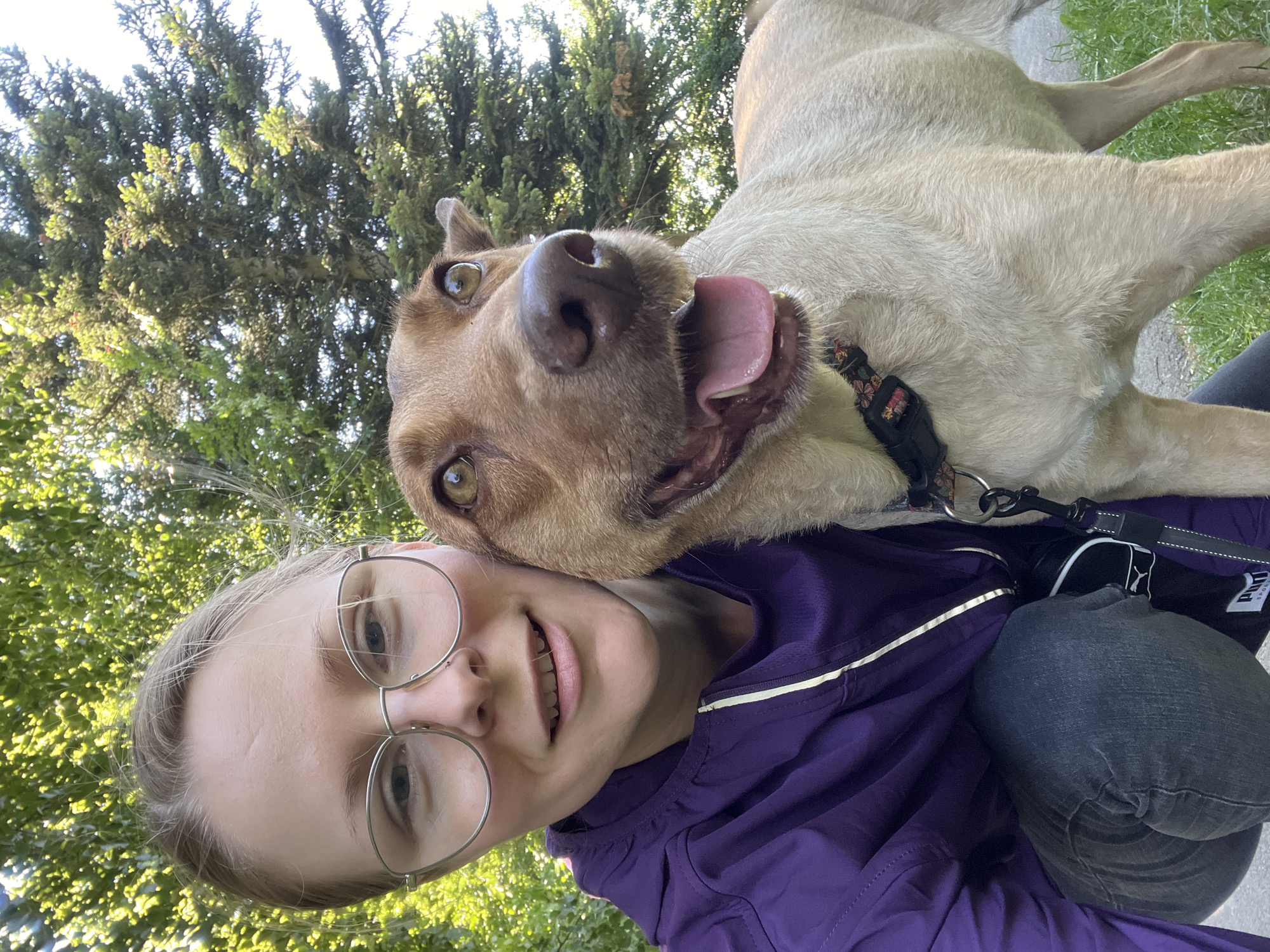 In Saint-Vith, Belgium, a woman wearing glasses and a purple jacket is captured in a selfie with her light-colored dog. The dog, with its mouth open and tongue out, is looking directly at the camera, its ears slightly raised. The background features lush green trees and foliage, suggesting an outdoor setting. The woman is crouching down, her arm around the dog, creating an intimate and playful moment between the two subjects. The lighting suggests it is daytime, possibly late afternoon given the warm tones.