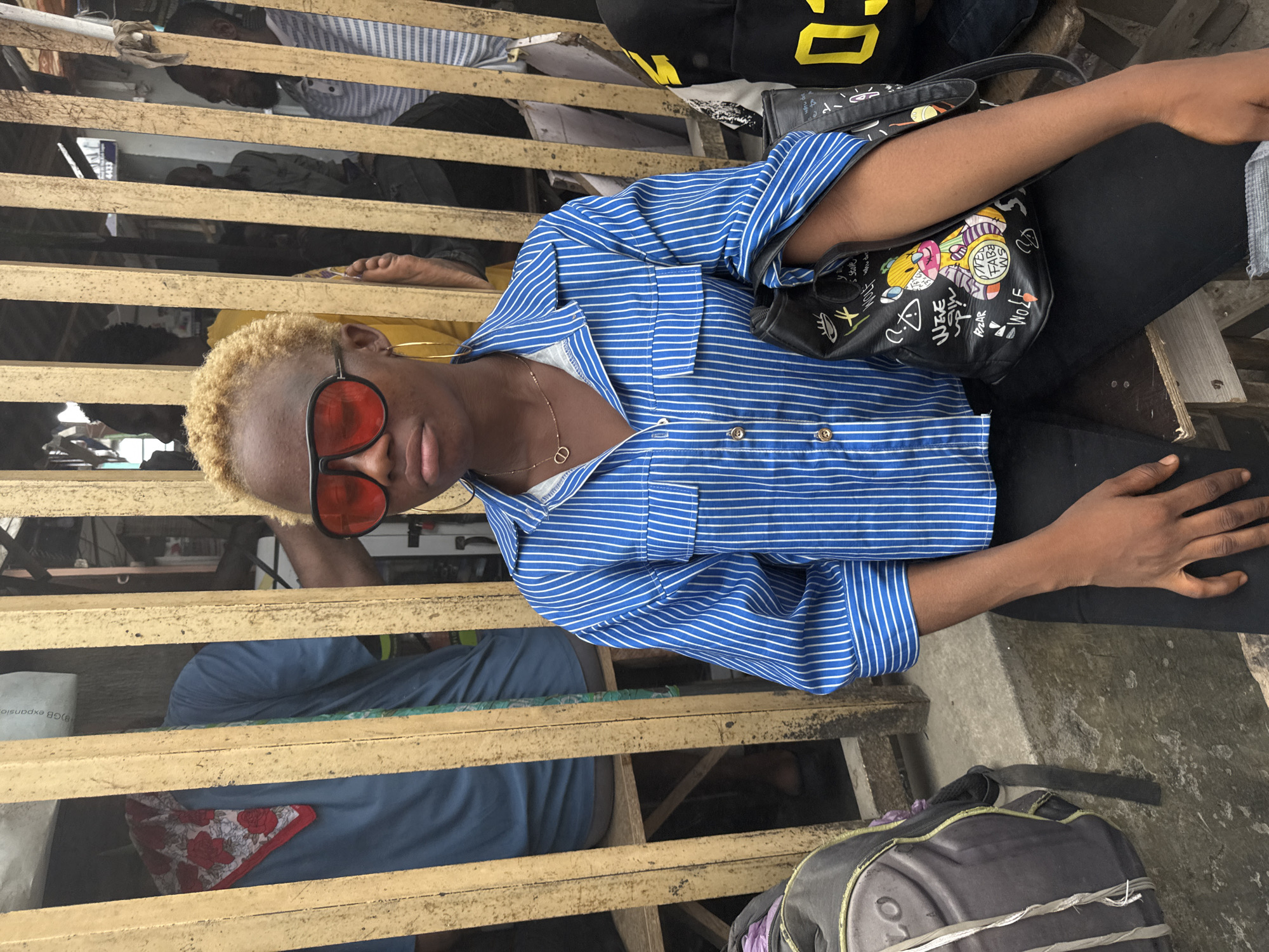 A person with bleached blond hair and red sunglasses sits near a wooden structure. The person wears a blue striped shirt, black pants, and a gold necklace. A black handbag with drawings is on their lap. A gray backpack is on the ground next to them. The scene appears to be outdoors, possibly a market or public area in Lagos, Nigeria, based on the architectural details of the wooden structure.