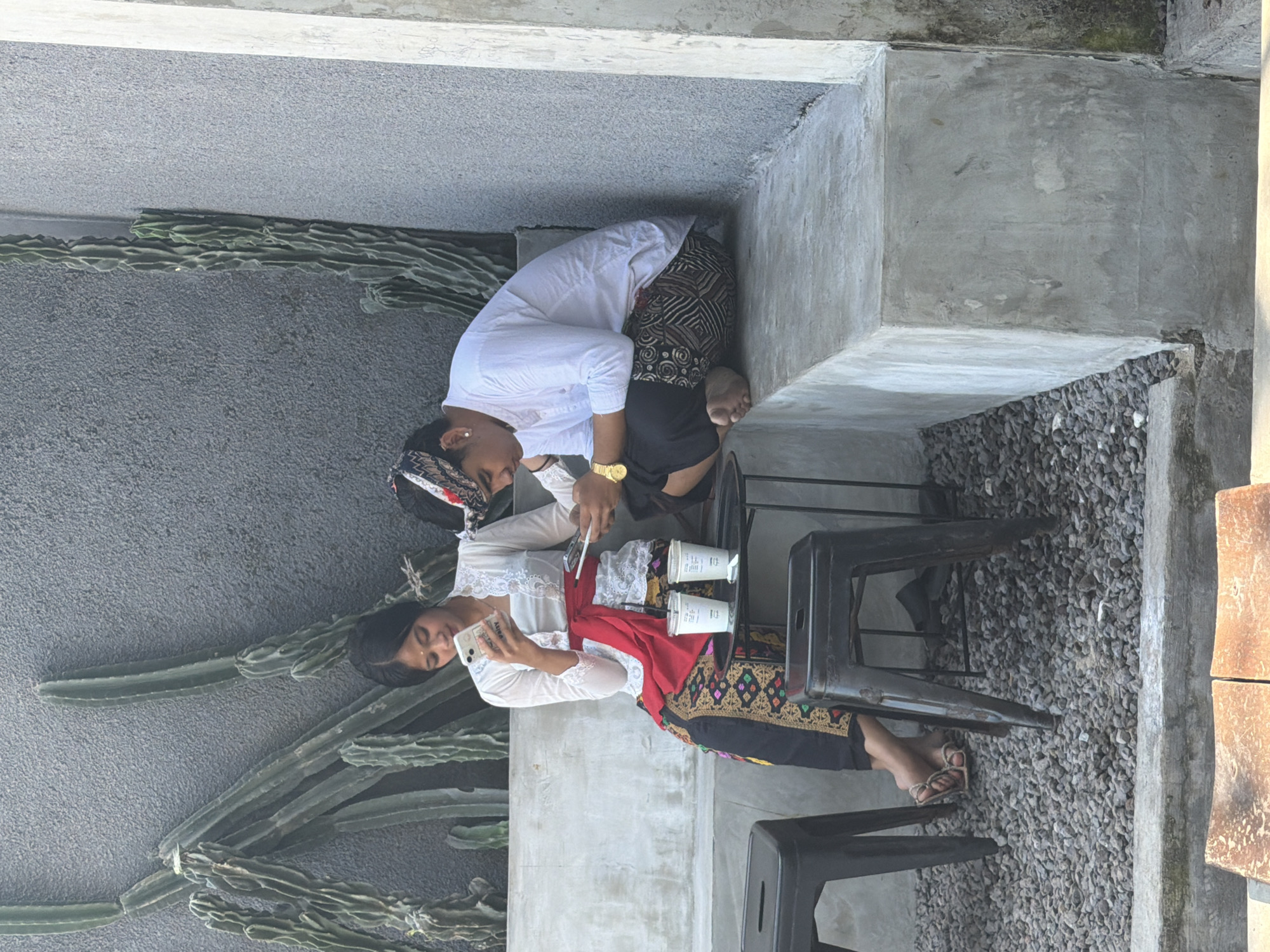Two women are sitting outdoors, near cacti and gray stone, potentially in Kuta, Indonesia, based on cultural clothing. One woman in a white top and patterned bottom sits while another adjusts her hair, also in a white top and patterned bottom with red accents. They are near two coffee cups, with a smartphone visible in the hands of one of the women. The setting is a seating area with black metal chairs and small gray rocks.