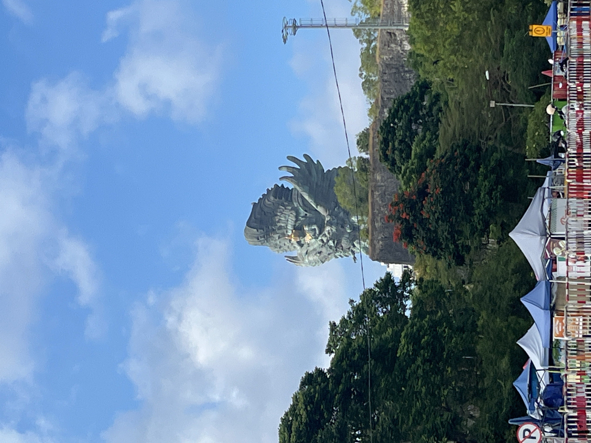 The image depicts the Garuda Wisnu Kencana Cultural Park in Kuta Selatan, Indonesia. In the background, a colossal bronze statue of Vishnu riding on Garuda dominates the skyline, reaching towards a bright blue sky with scattered white clouds. The statue is depicted with intricate details, showcasing Vishnu seated on the mythical bird, with outstretched wings. The statue is perched on a rocky outcrop, surrounded by lush green trees and foliage, some of which display vibrant red flowers. In the foreground, a series of tents and stalls are set up, suggesting a marketplace or event. Several people are visible near the tents, and some motorbikes are parked behind a barrier. A "P" sign for parking is also visible to the left. A tall communication tower with a light fixture stands to the right of the statue. The overall scene captures a blend of cultural heritage and public gathering within a tropical environment.