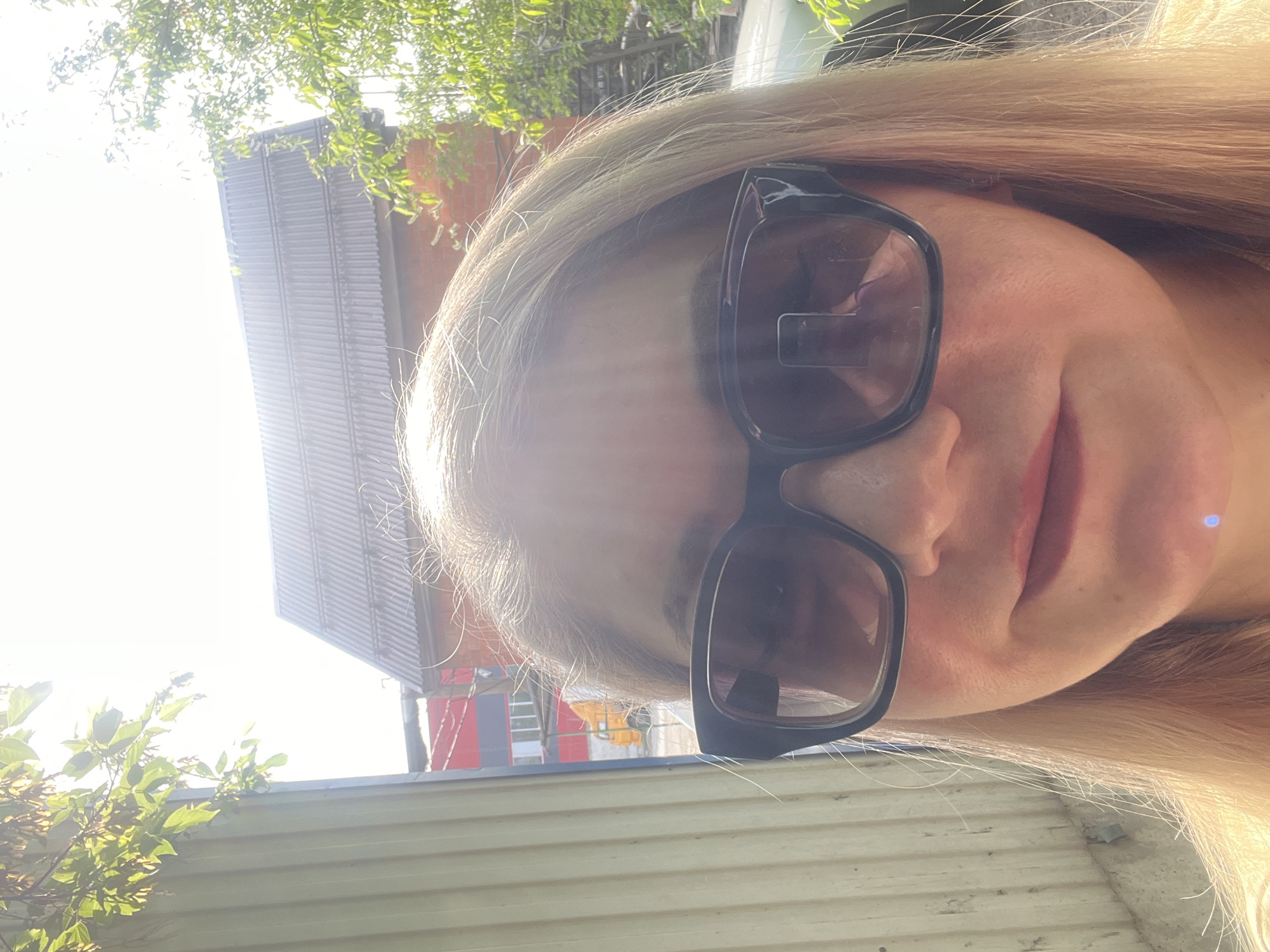 A selfie of a fair-skinned woman with long, blonde hair and dark-framed sunglasses. The background shows part of a building with a metal roof and a red brick exterior. There's also green foliage. The image was taken outdoors in bright sunlight. It is not possible to verify the exact location as Izhevsk, Russia from the image alone.