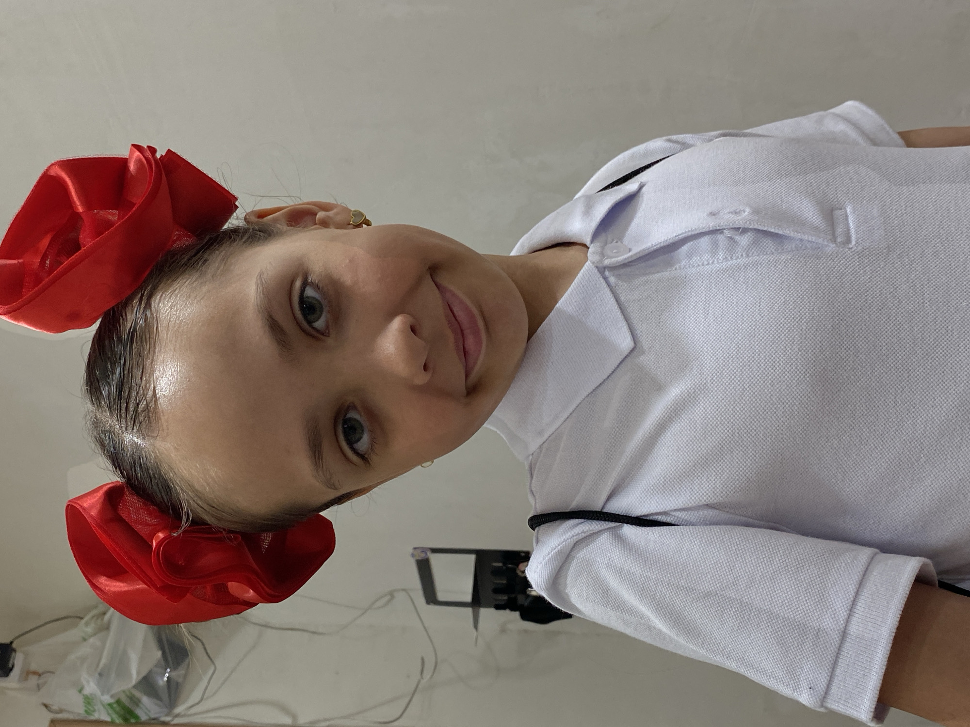 A young girl is shown in a portrait-style image. She has light skin, light-colored eyes, and wears a white polo shirt with a collar and buttons. Her hair is pulled back and adorned with two large, bright red satin bows. The background is a plain, light-colored wall. There are some indiscernible objects on a surface below the frame, along with some wires and possibly a bag. The image's location is unknown.