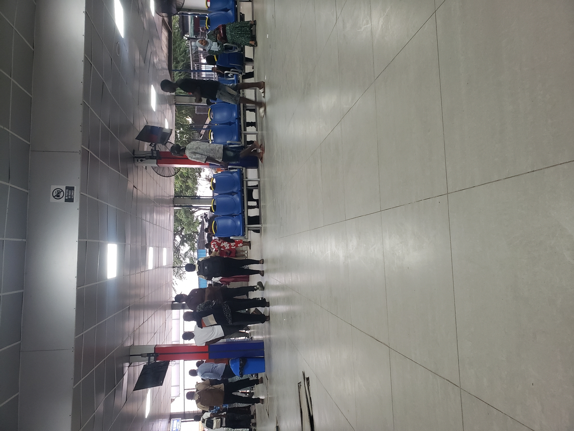 The image depicts an indoor waiting area, likely a bus terminal or train station, in Dar es Salaam, Tanzania. The space features a tiled floor and a suspended ceiling with fluorescent lighting. Rows of blue plastic chairs are arranged for waiting passengers. Several people are present, some standing and others seated. One man in the foreground is wearing a black t-shirt and denim shorts, walking on the tiled floor. To his left, another man in a striped t-shirt and shorts is also walking. In the background, individuals are seen waiting, with some carrying bags. A sign indicating "NO PHOTOS OR VIDEO" is visible on the ceiling. The environment appears to be public and functional, with a noticeable amount of space for people to gather.