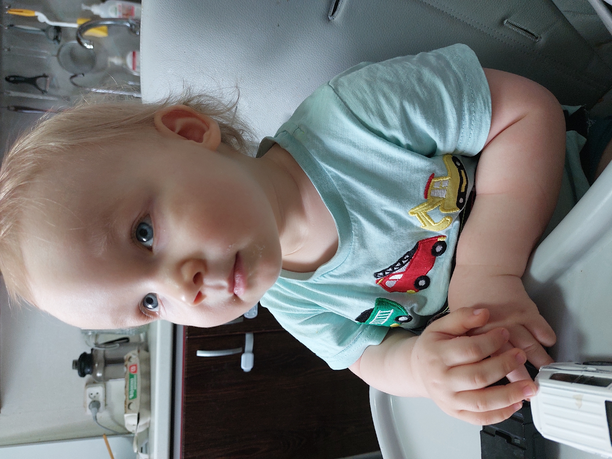 A fair-skinned, light-haired toddler wearing a light blue t-shirt with car graphics is sitting in a high chair. The child has food residue around their mouth. The child's hands are clasped together, and they appear to be playing with a toy. The background is a kitchen environment, with kitchen utensils and appliances visible. There is no explicit information within the image to determine the location is Chaikovsky, Russia.