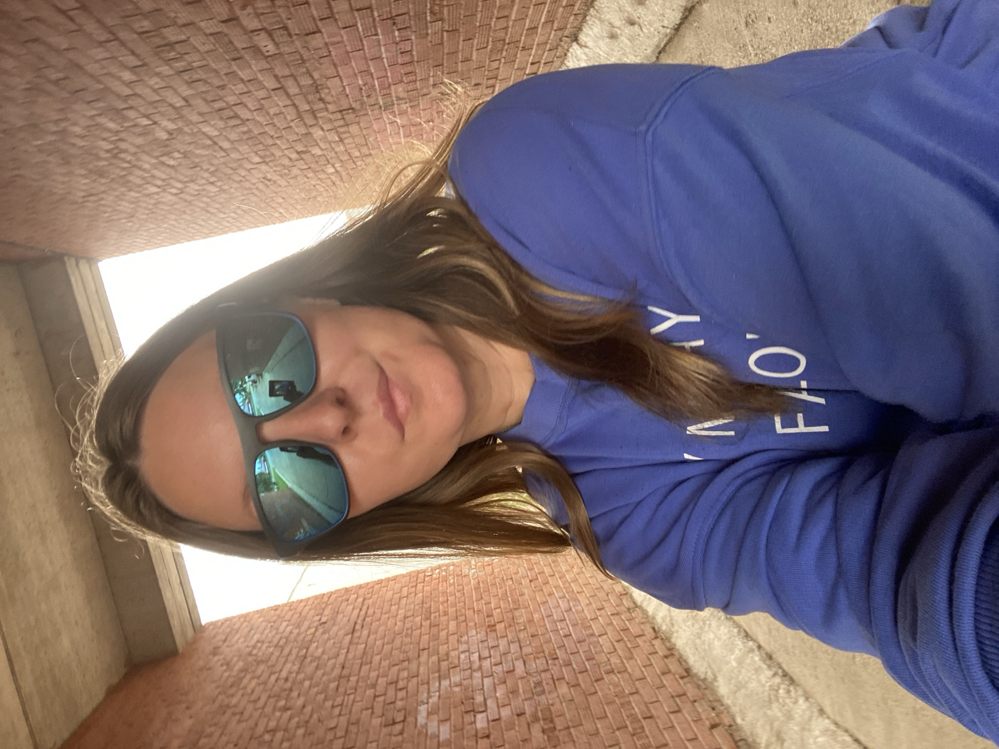 A woman, wearing sunglasses and a blue sweatshirt, is taking a selfie. The sweatshirt has white text on it, but it is partially obscured. She is standing in a narrow walkway with brick walls on either side, and a concrete ceiling overhead. The walkway appears to be an underpass or tunnel. Sunlight streams in from the end of the walkway. The background is blurred and out of focus. The scene is likely an outdoor location in the city of Noviy, Russia.