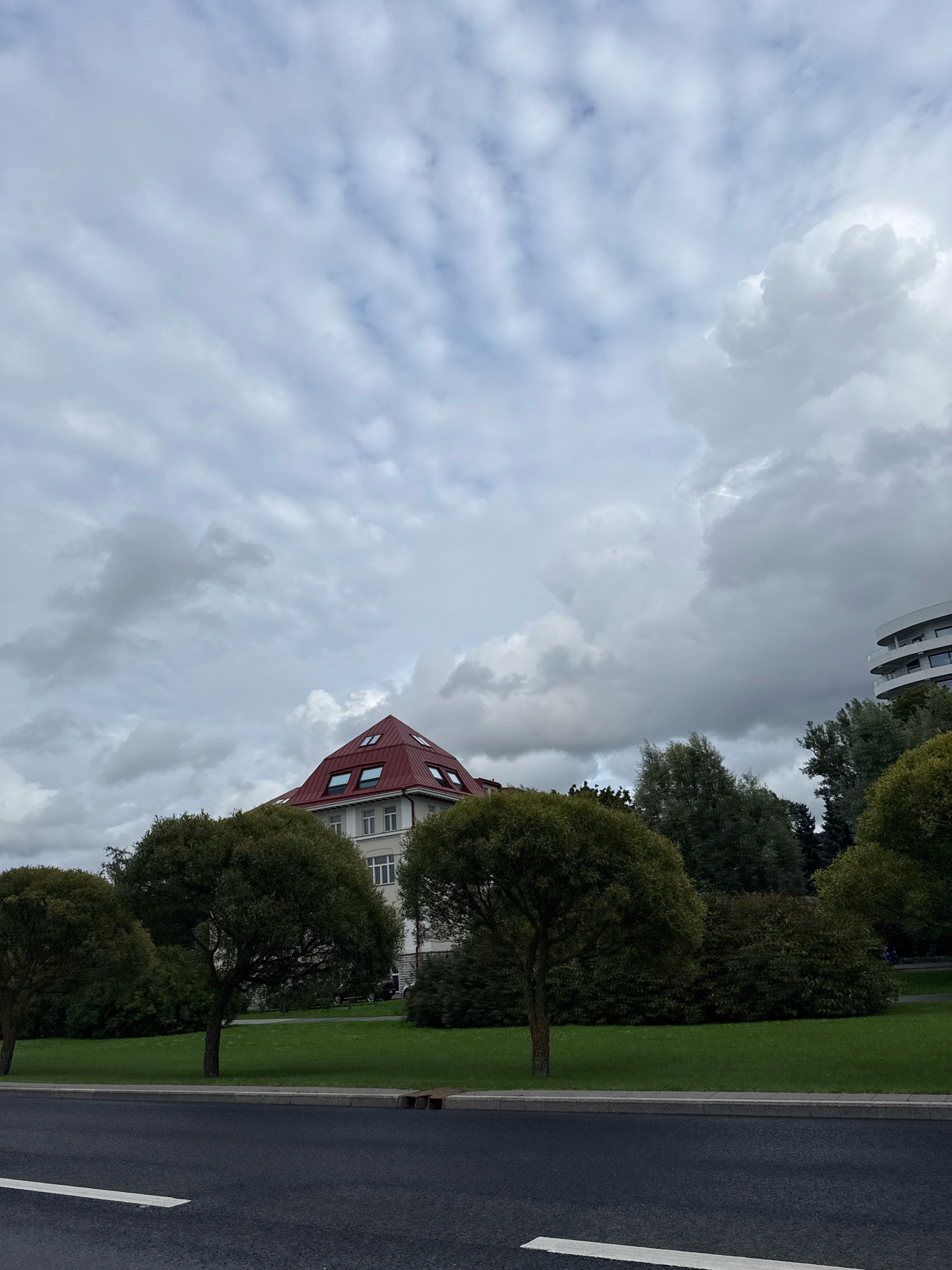 The image depicts a cloudy day in Tallinn, Estonia. In the foreground, a dark asphalt road with a white line runs horizontally across the bottom of the frame. Behind the road, a strip of grass and a sidewalk are visible, bordered by a row of lush green trees and bushes. In the mid-ground, a building with a distinctive red, pyramid-shaped roof and multiple windows is partially obscured by the trees. To the right of this building, another modern building with a curved facade and balconies can be seen peeking above the foliage. The sky is filled with a dramatic arrangement of grey and white clouds, creating a moody atmosphere.