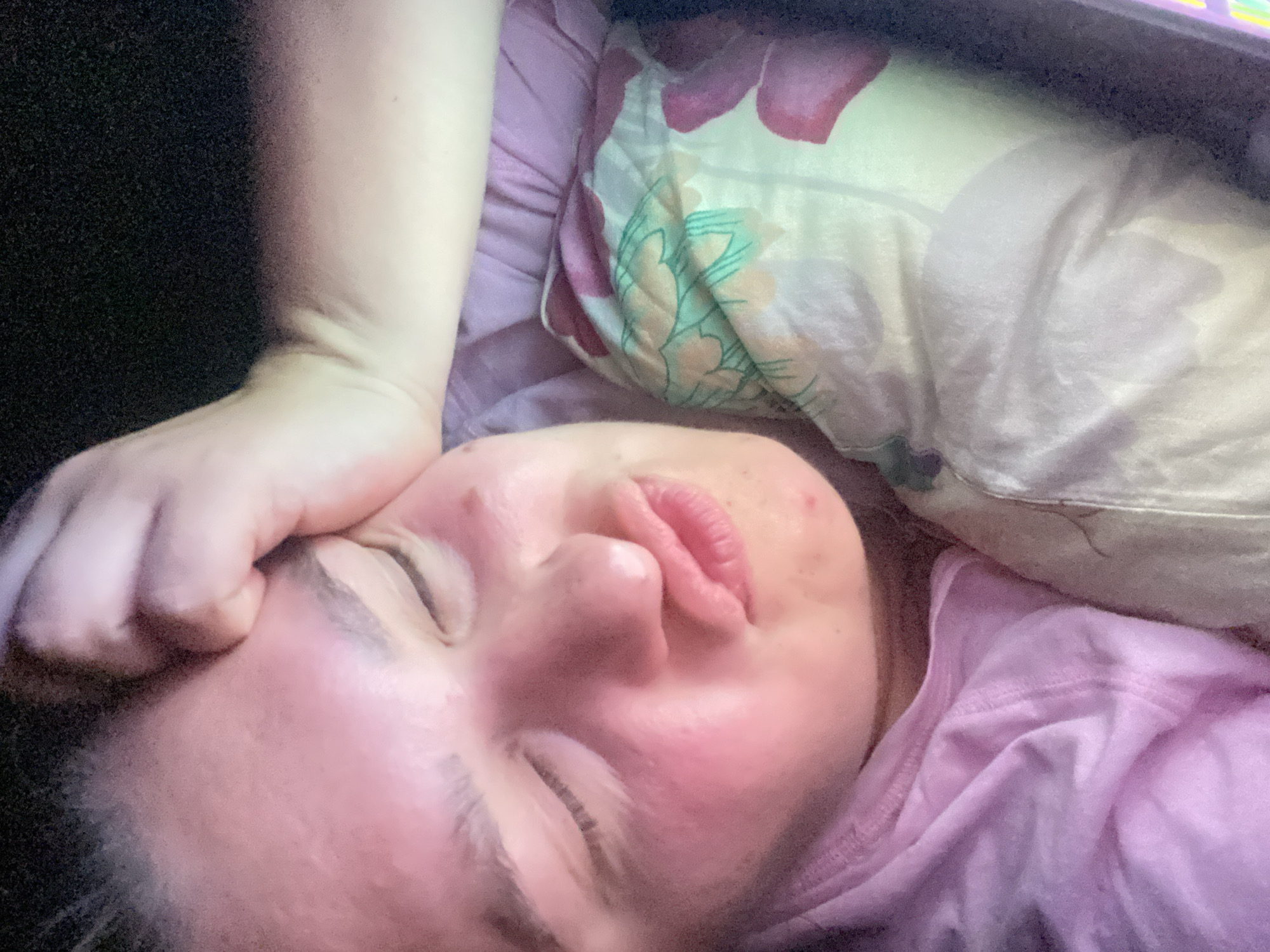 The image shows a close-up, low-angle selfie of a person lying down. The person's face is partially visible, with eyes closed and lips pursed. They are resting their forehead on their hand. The person is wearing a pink t-shirt and is lying on a pillow with a floral pattern. The background is dark, suggesting a dimly lit indoor environment. There are no discernible landmarks or contextual clues to indicate a specific location within the city of Perm, Russia.