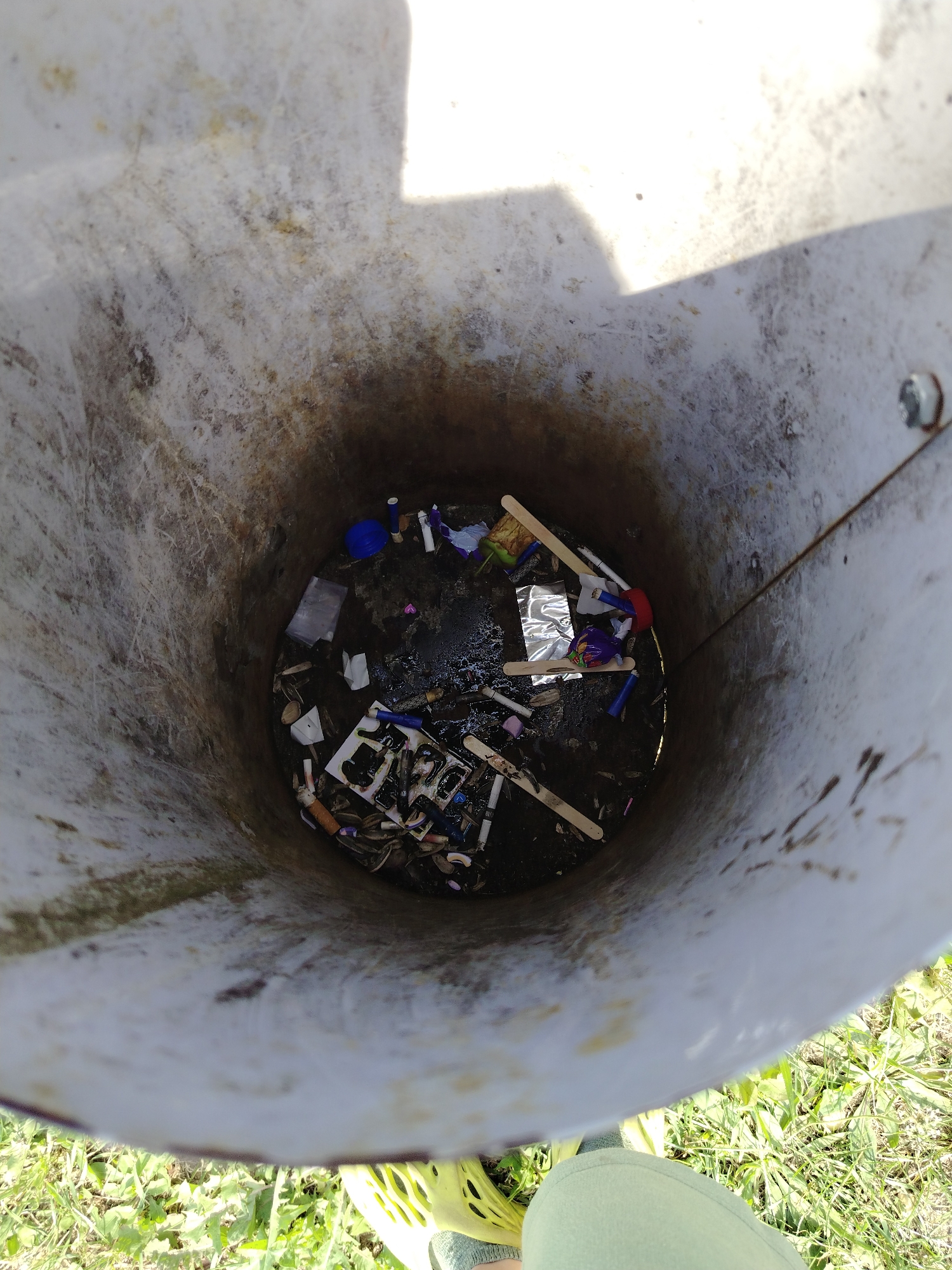 A trash can is overflowing with garbage. The trash can is made of a white plastic material and is dirty and stained. The garbage inside consists of various items, including plastic bottles, wrappers, food scraps, and cigarette butts. Some of the garbage is wet, suggesting that it may have rained recently or that the trash can is collecting liquid waste. The trash can is located outdoors, on a patch of grass. The lighting suggests that it is daytime.