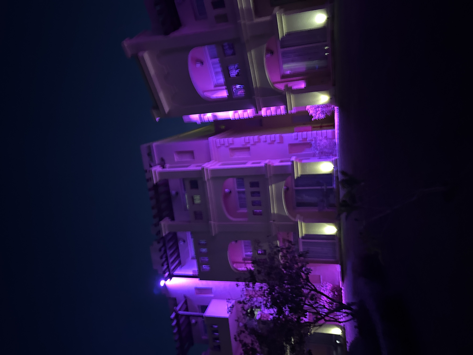 This is a photograph of a modern, multi-story building illuminated by purple and yellow lights at night. The building appears to be a resort or hotel, with balconies and arched doorways. A tree with purple-hued foliage is visible in the foreground. The sky above is dark blue, indicating it is night. The image is slightly blurry due to low light conditions. The location is specified as an unknown city in Egypt.