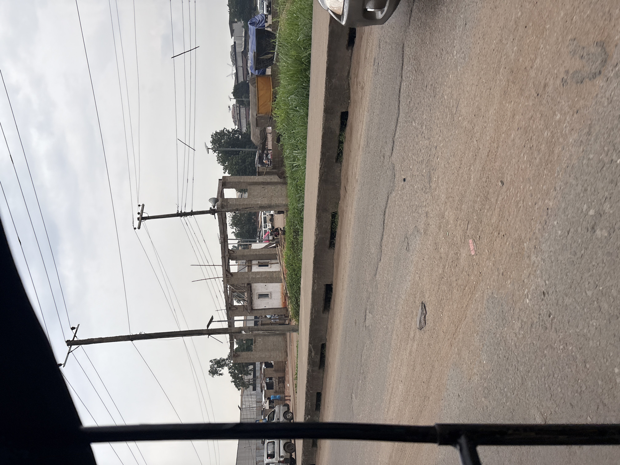 A medium shot, taken from inside a moving vehicle, shows a street scene in Onitsha, Nigeria. The foreground is dominated by the road surface, which appears to be asphalt with some patches of dirt and debris. A concrete curb with drainage openings runs along the side of the road. Beyond the curb, there is an area of overgrown green grass and a concrete structure that appears to be under construction, with visible rebar and cinder blocks. Several makeshift stalls covered with tarpaulins are set up behind the construction area. In the background, there are more buildings and some vehicles, including vans and cars. Overhead, there are multiple electrical wires supported by wooden poles. The sky is overcast. A person wearing a blue shirt and dark shorts is standing near the construction area, and another person is partially visible to their left. A white van is parked to the left of the frame.