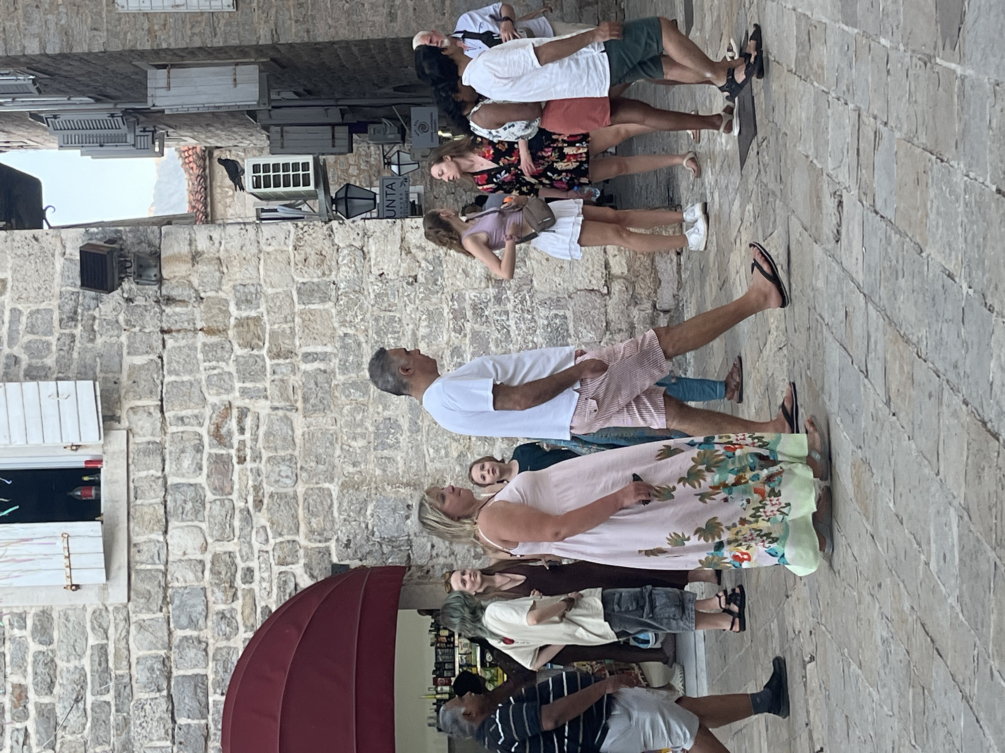 The image depicts a street scene in the historic Old Town of Budva, Montenegro. The architecture features rough, stone walls and traditional shutters. A group of people are walking on a cobblestone street.
In the foreground, a man in a white t-shirt and striped shorts is walking away from the camera. To his left, a woman in a light pink maxi dress adorned with a tropical print walks towards the camera. Behind her, a young boy with short brown hair looks towards the left. Further left, a man in a striped t-shirt and white shorts walks towards the camera.
To the right of the main couple, a group of women are interacting. One woman in a light purple tank top and white skirt is facing a woman in a black floral dress. Another woman in a white top with a floral pattern and an orange skirt is partially visible. A man with white hair and a light blue shirt is in the background on the far right.
In the background, a red awning shades a shop entrance. Above the people and to the left of the image, a stone wall with a window is visible, and inside the window, a red bottle and a yellow can are on a sill. On the wall to the right, an air conditioning unit is mounted, and a pigeon is perched on it. Street lamps are also visible mounted on the wall. The overall impression is a lively, sunlit street with tourists and locals enjoying the atmosphere.
