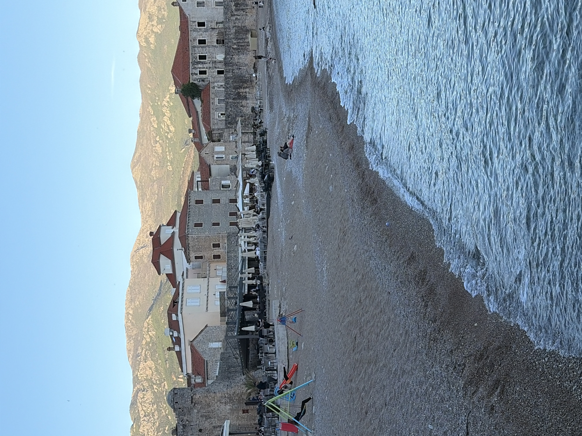 This image captures a vibrant coastal scene in Budva, Montenegro, likely during the late afternoon or early evening. The view is from slightly above, looking down towards a pebble beach, the sea, and a historical town nestled against a mountain range.
On the right, gentle waves of the Adriatic Sea break softly onto the shore, exhibiting a pattern of frothy white lines. The beach, composed of small pebbles and sand, stretches along the coastline.
In the midground, several people are visible. One individual, dressed in red, is sitting directly on the beach near the water's edge, while others are scattered further back. Towards the left of the beach, a children's play area with colorful swings and a small slide is present. Adjacent to this, an outdoor cafe or restaurant area with numerous tables, chairs, and umbrellas is set up, bustling with what appear to be patrons.
The background is dominated by ancient-looking stone buildings with distinctive red-tiled roofs, characteristic of traditional Mediterranean architecture. These multi-story structures line the shore, some featuring visible windows and balconies. Behind these buildings, a majestic, rugged mountain range rises steeply, its rocky, sparsely vegetated slopes illuminated by the soft, warm light of the setting sun. The sky above is clear and a pale blue, indicating pleasant weather.
The overall atmosphere is one of leisure and tranquility, depicting a popular tourist destination combining natural beauty with cultural heritage. There is no visible text in the image.