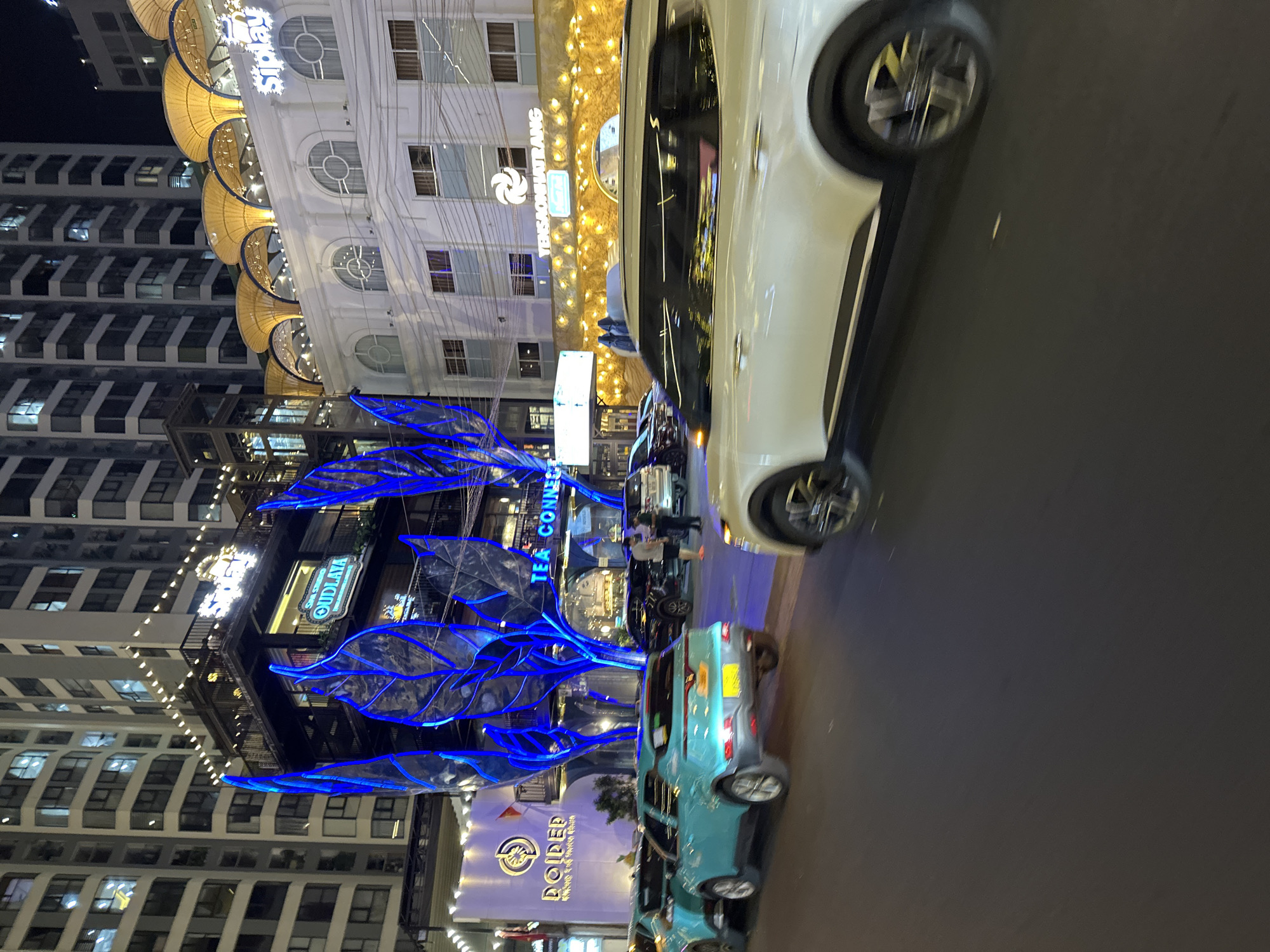 The image depicts a night scene in Nha Trang, Vietnam, likely on a busy street. In the foreground, a white SUV is prominently displayed, its motion blurred due to camera shake or movement. To the left, several cars are parked or moving, including two teal-colored vehicles, one of which is closer to the viewer and appears to be a hatchback. Behind the vehicles, a multi-story building with numerous lit windows dominates the background. The building features illuminated signage, with "Sip Lay" visible in large white letters. Another sign with a circular logo and the text "DOIPED" is also present, along with "TEA CONNECTION" written in white lettering. The architecture suggests commercial establishments or residential units. The street is paved and appears to be asphalt. The overall atmosphere is illuminated by artificial lights from the buildings and vehicles, creating a vibrant, urban night setting.