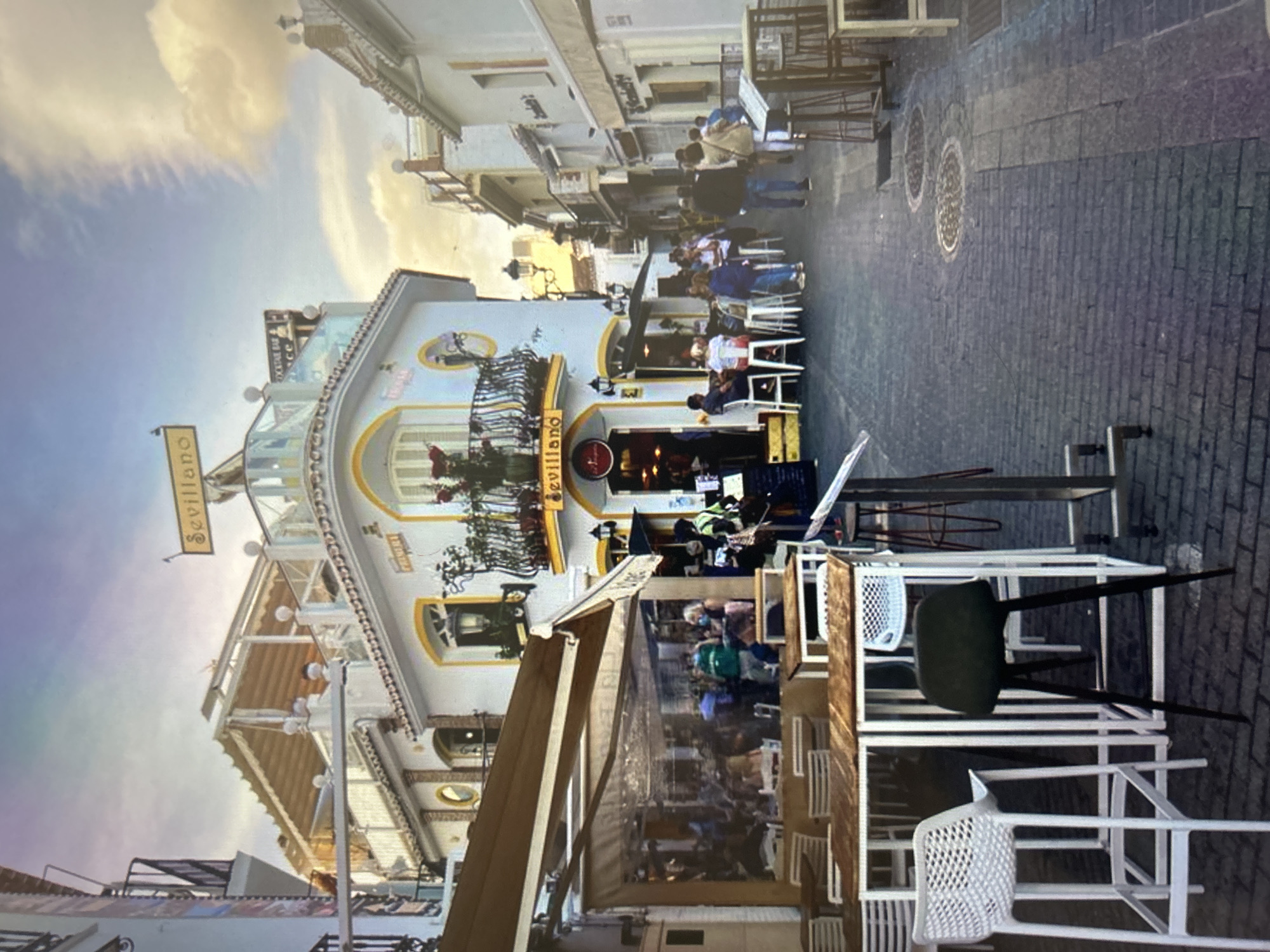 The image captures a lively daytime street scene, likely in a bustling European town or city. The central focus is a multi-story white building with yellow trim and balconies, housing a restaurant or bar. A prominent sign hanging from the building reads "Sevillano." Below this sign, other text like "COCKTAIL BAR" and potentially "Dance" is partially visible on an upper glass-enclosed section.
Numerous people are present, seated at outdoor tables and chairs on the paved street, or walking. They appear to be patrons enjoying al fresco dining or socializing. The street itself is paved with grey and brown brick-like material, and two circular manhole covers are visible. The sky is bright with some clouds, suggesting pleasant weather, likely late afternoon or early evening given the warm light. The overall atmosphere appears relaxed and social, with individuals enjoying the outdoor setting. The architecture, including the balconies and narrow street, along with the "Sevillano" sign, strongly suggests a Spanish-influenced location, possibly in Seville.