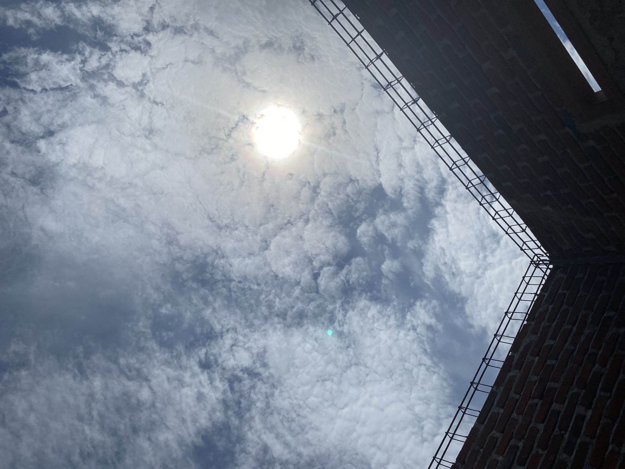 A low-angle, full shot shows a partly constructed brick building against a sky filled with scattered clouds and the bright sun. The sun is in the upper right quadrant of the image, a bright white orb with visible rays, partially obscured by clouds. The sky is a mix of blue and white, with wispy clouds dominating the upper portion. The brick walls of the building, made of dark red bricks, rise from the bottom of the frame, forming an interior corner. Rebar, bent into a cage-like structure, runs along the top edge of the brick walls. The building appears unfinished, suggesting it is under construction. There are no people visible in the image. The environment is outdoors, under a daytime sky.