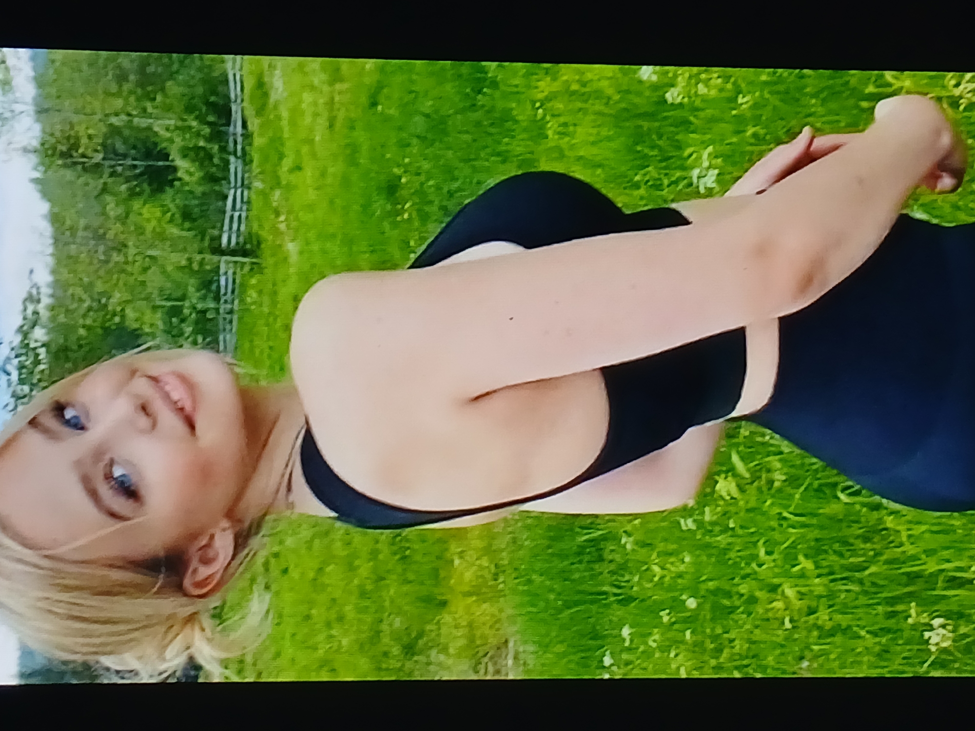 The image shows a young woman with blonde hair pulled back, wearing a black sports bra and leggings, posing outdoors in a grassy field with trees and a wooden fence in the background. She is looking over her left shoulder at the camera with a smile. The lighting suggests it is daytime. The image quality is moderate. No specific location in Dnipro, Ukraine can be identified from the provided visual information.
