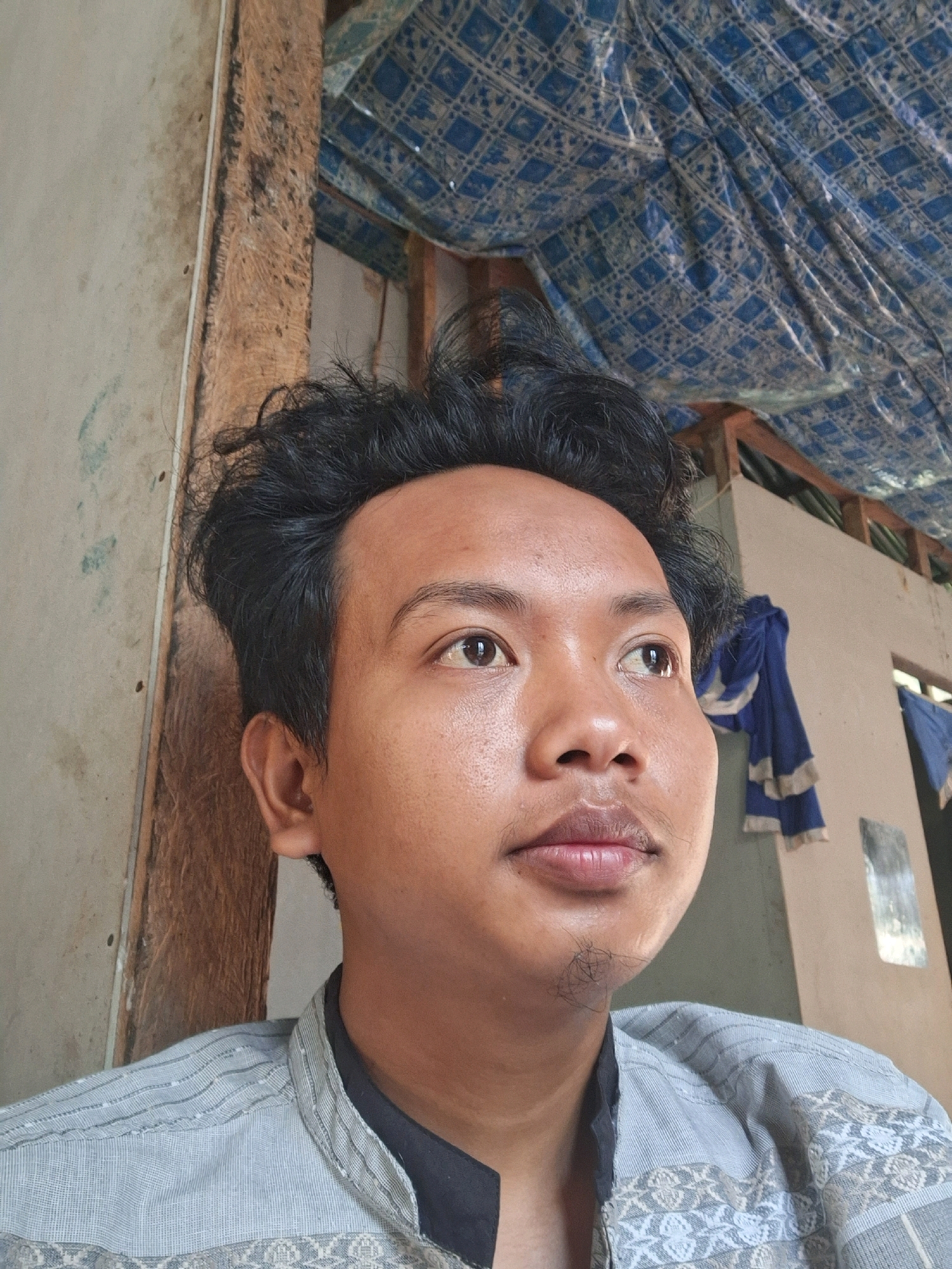 A medium-shot portrait shows a young man looking to his right, his face filling most of the frame. He has dark, unkempt hair and is wearing a light gray collared shirt over a dark garment. The man has visible stubble on his chin. The background appears to be an interior space with a rough, wooden beam to the left and a blue patterned fabric overhead, suggesting a simple structure or shelter. The lighting is natural and somewhat diffuse. The image is likely a selfie taken in a residential or communal area within Air Tawar, Indonesia, given the context provided.