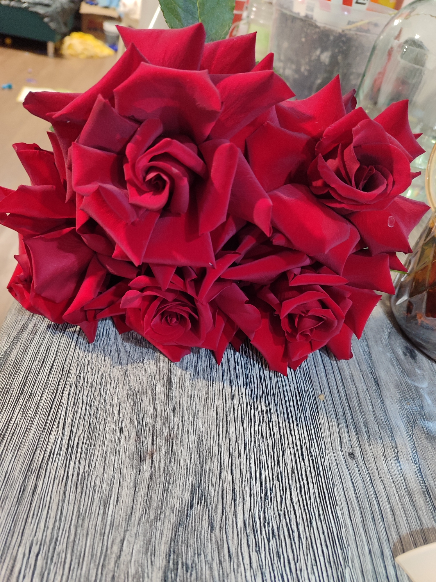 This media file features a close-up, high-angle shot of a vibrant bouquet of deep red roses. The roses are the central subject, clustered tightly together, displaying their velvety petals and intricate, spiraling centers. Several individual blooms are visible, ranging from tightly furled buds to more open flowers. Some petals exhibit small, clear water droplets, suggesting freshness.
The bouquet rests on a light grey or weathered wooden surface, which occupies the lower half of the frame. This surface has a distinct, pronounced vertical grain pattern, providing a textured contrast to the soft petals of the roses.
The background is intentionally blurred due to a shallow depth of field, drawing focus to the flowers. Despite the blur, an indoor setting is evident, with glimpses of household items. On the left side of the background, a portion of a blue-green piece of furniture or fabric is visible, along with a yellow object. To the right and center-back, several glass containers can be vaguely discerned; one appears to hold dark soil or a similar substance, while another is a clear, patterned glass vessel, possibly a vase or bottle.
There are no people visible, and no specific activity is taking place beyond the static arrangement of the flowers. The image captures a moment of natural beauty in an indoor setting. No visible text is present within the image. The location context for this media file is Izhevsk, Russia.