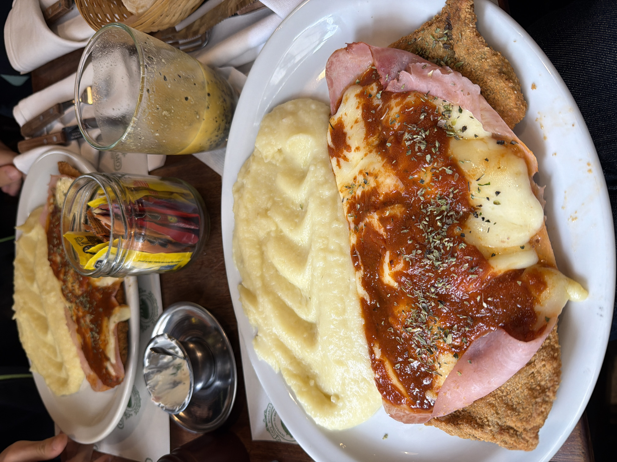 The image captures a meal, likely in a restaurant setting, in Buenos Aires, Argentina. Two plates are visible, each containing a serving of mashed potatoes and what appears to be a Milanese sandwich (a breaded meat cutlet). The sandwich is topped with ham, melted cheese, and a tomato-based sauce, sprinkled with dried herbs. A tall glass of a yellow liquid, possibly a juice or smoothie, sits near the plates. A jar filled with packets of sweeteners and a small saucer containing a white substance, possibly butter or cream cheese, are also present on the wooden table. The scene suggests a casual dining experience, possibly at a local café.