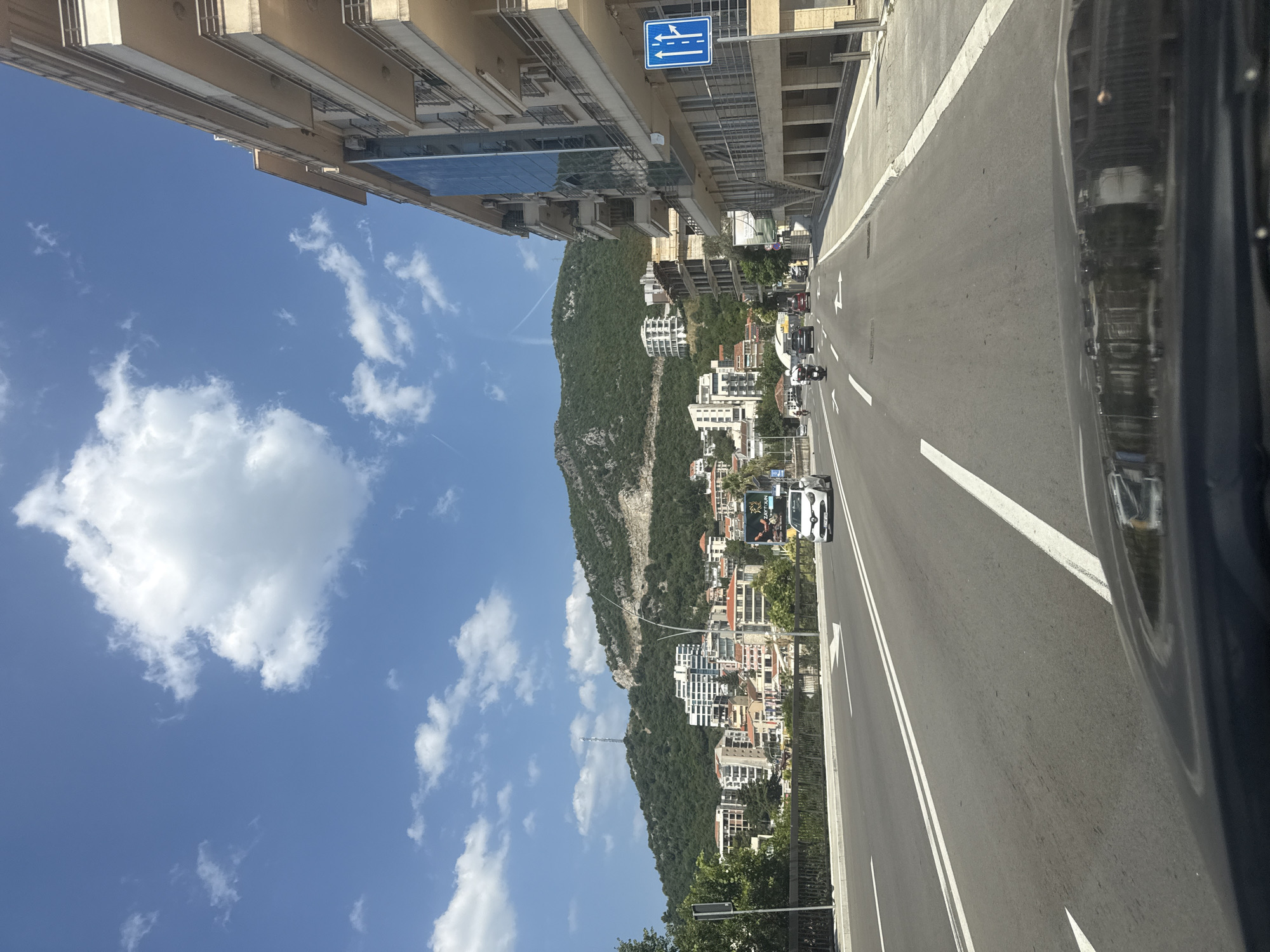The image depicts a street scene in Boreti, Montenegro, on a clear, sunny day with scattered white clouds in a blue sky. A multi-lane asphalt road runs through the center of the frame. On the right side of the road, a modern, multi-story building with balconies and glass elements is visible. A blue traffic sign indicating lane usage is mounted on a pole in front of the building.
The road has white lane markings and directional arrows painted on its surface. Several vehicles are present: a white car in the right lane, a bus in the left lane, and a motorcycle further ahead in the left lane. In the distance, a lush green, mountainous landscape dominates the background, dotted with various buildings, including hotels and residential structures. Trees and vegetation are also visible on the hillsides. The image is captured from a low angle, likely from within a vehicle, as the hood of a car is visible in the foreground.
