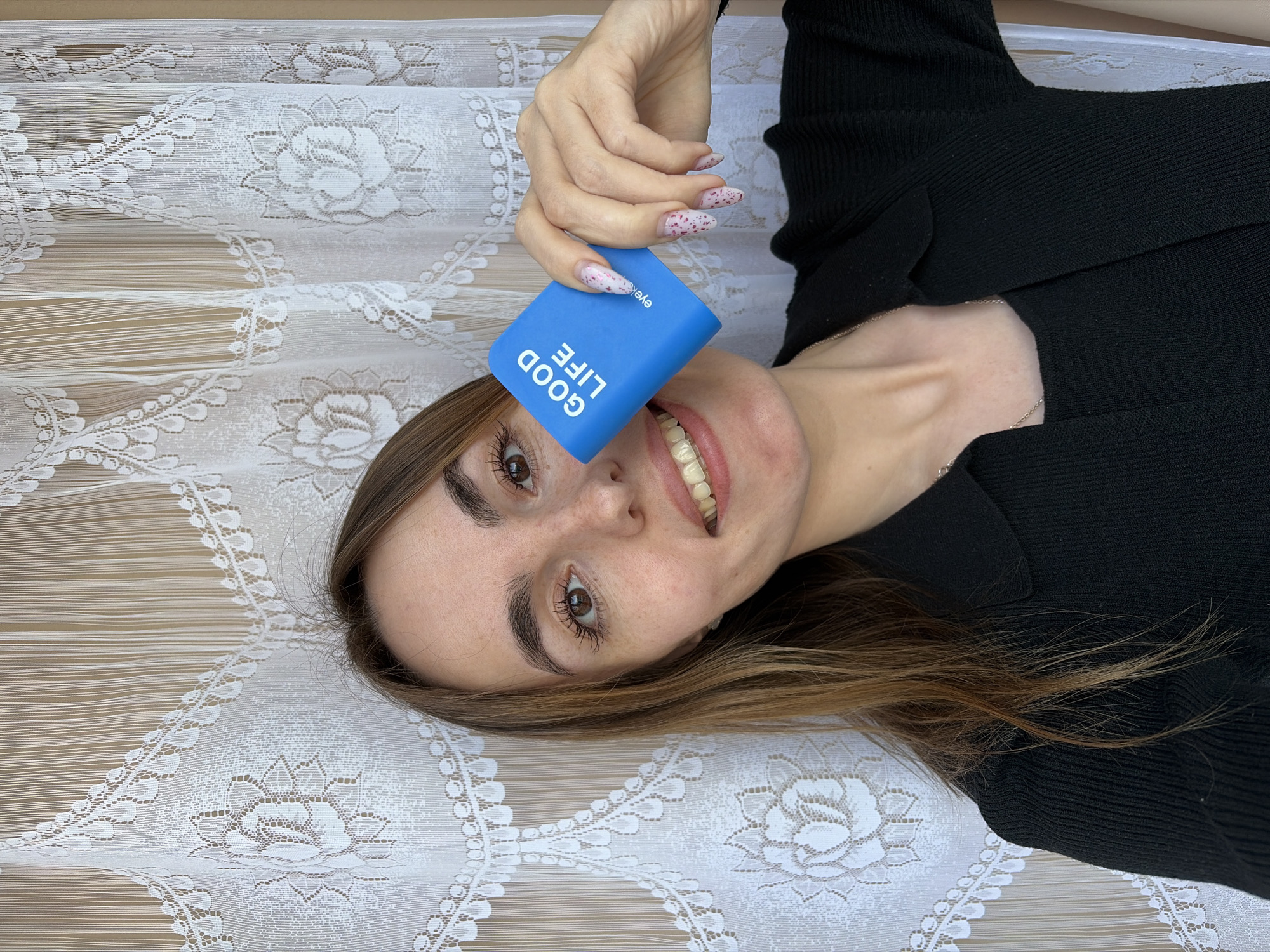 A fair-skinned, brown-haired woman with hazel eyes, wearing a black knit top and a thin silver necklace, lies supine on a surface covered with white floral lace fabric. She is holding a blue rectangular object labeled "GOOD LIFE" near her face. The woman's fingernails are manicured with a white and pink speckled design. The image does not indicate a location in the city of Izhevsk, Russia.