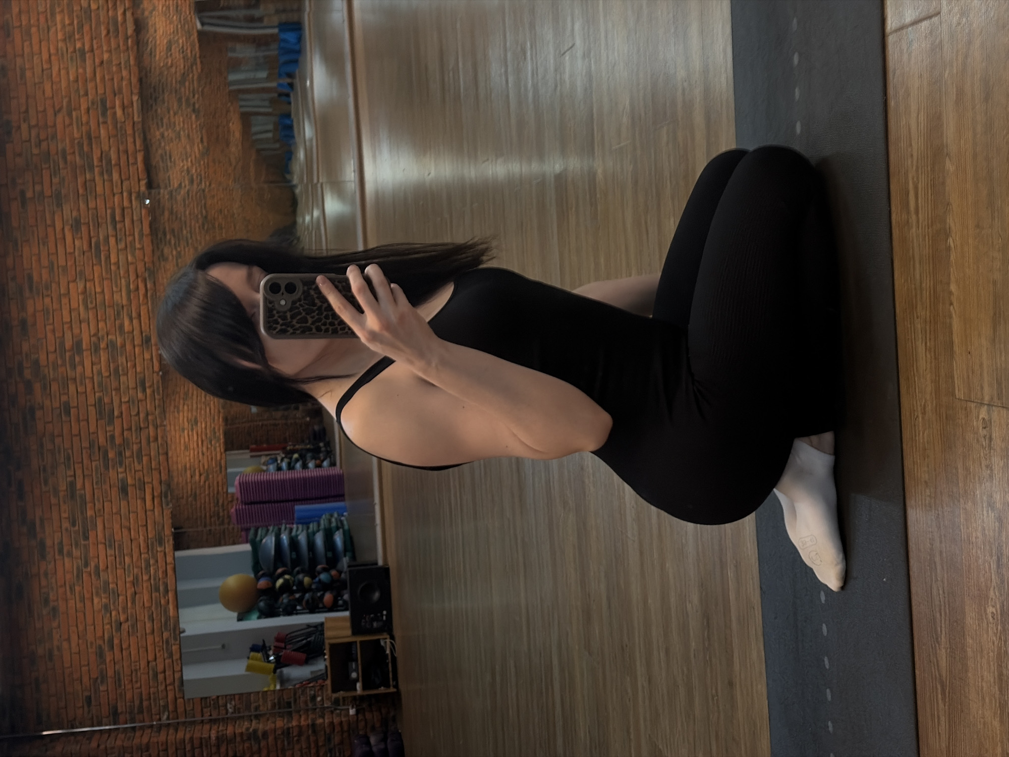 A person is seated in a gym or studio. The individual has dark hair, is wearing a black bodysuit, and one white sock. They are holding a smartphone with a patterned case and taking a picture in front of a mirror, which reflects gym equipment. The floor is made of wood, and the person is positioned on a dark yoga mat. The location is suggested to be in Chaikovsky, Russia, although there is no direct evidence within the image to confirm this.