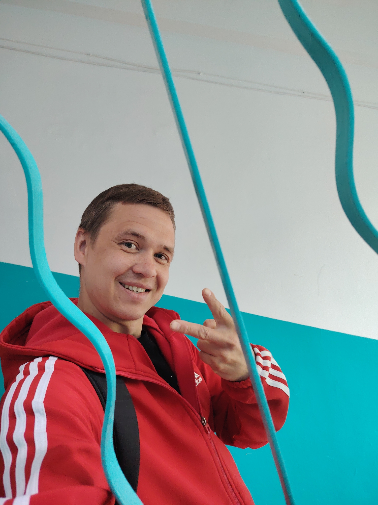 A man stands smiling in front of a blue railing. The man is wearing a red Adidas jacket with white stripes down the sleeves and a black shirt. He's making a hand gesture with his right hand. A glimpse of a black backpack strap is visible on his left shoulder. The railing is in front of a wall with a white top and a blue bottom section. The photo was taken in the city of Chaikovsky, Russia.